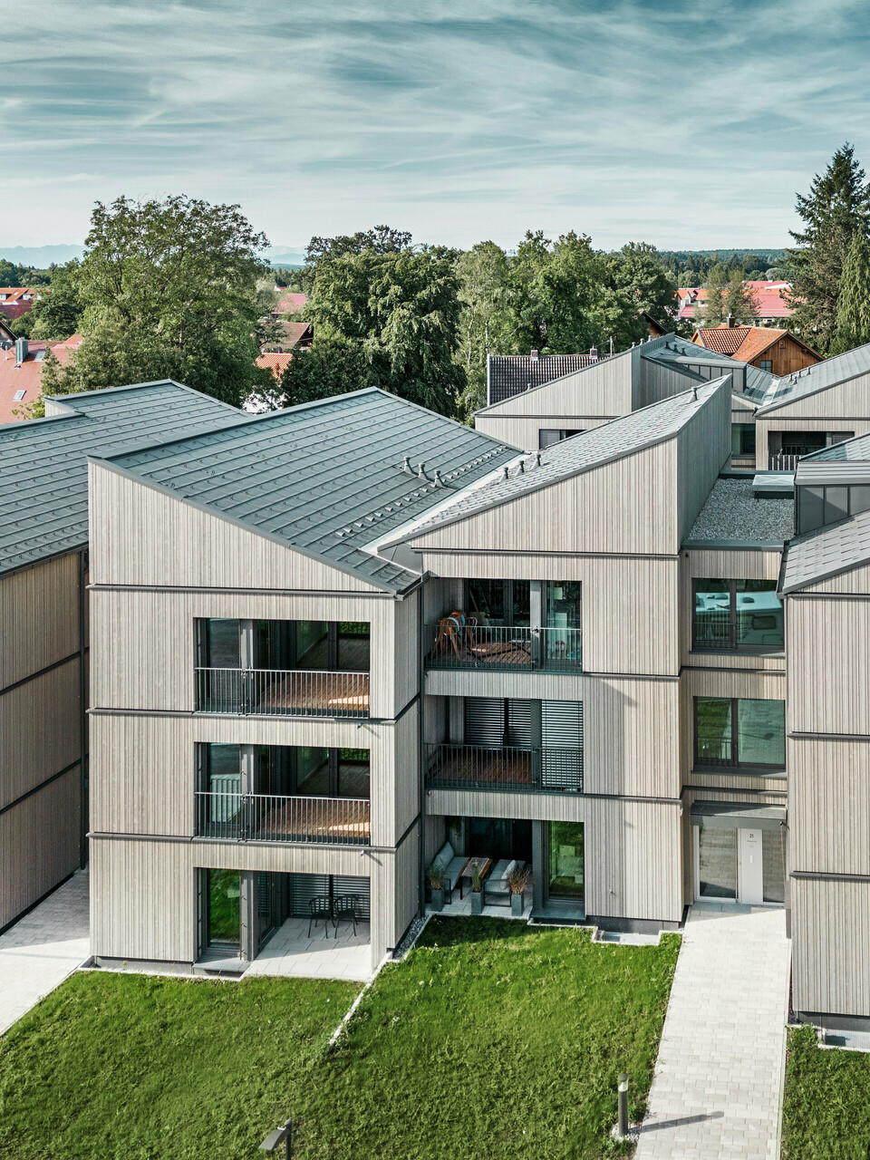 Luftaufnahme des modernen Mehrfamilienhauses am Schmucker-Areal in Utting am Ammersee, mit nachhaltiger Holzhybridbauweise und eleganten Holzfassaden. Die Gebäude sind mit langlebigen PREFA Dachplatten R.16 in P.10 Dunkelgrau ausgestattet. Diese innovative Architektur, geplant von WWA Architekten aus München, zeichnet sich durch klare Linien und harmonische Integration in die grüne Umgebung aus. Die großzügigen Balkone und großen Fenster sorgen für eine helle und freundliche Wohnatmosphäre, während die hochwertigen Materialien für Langlebigkeit und Wetterbeständigkeit sorgen.