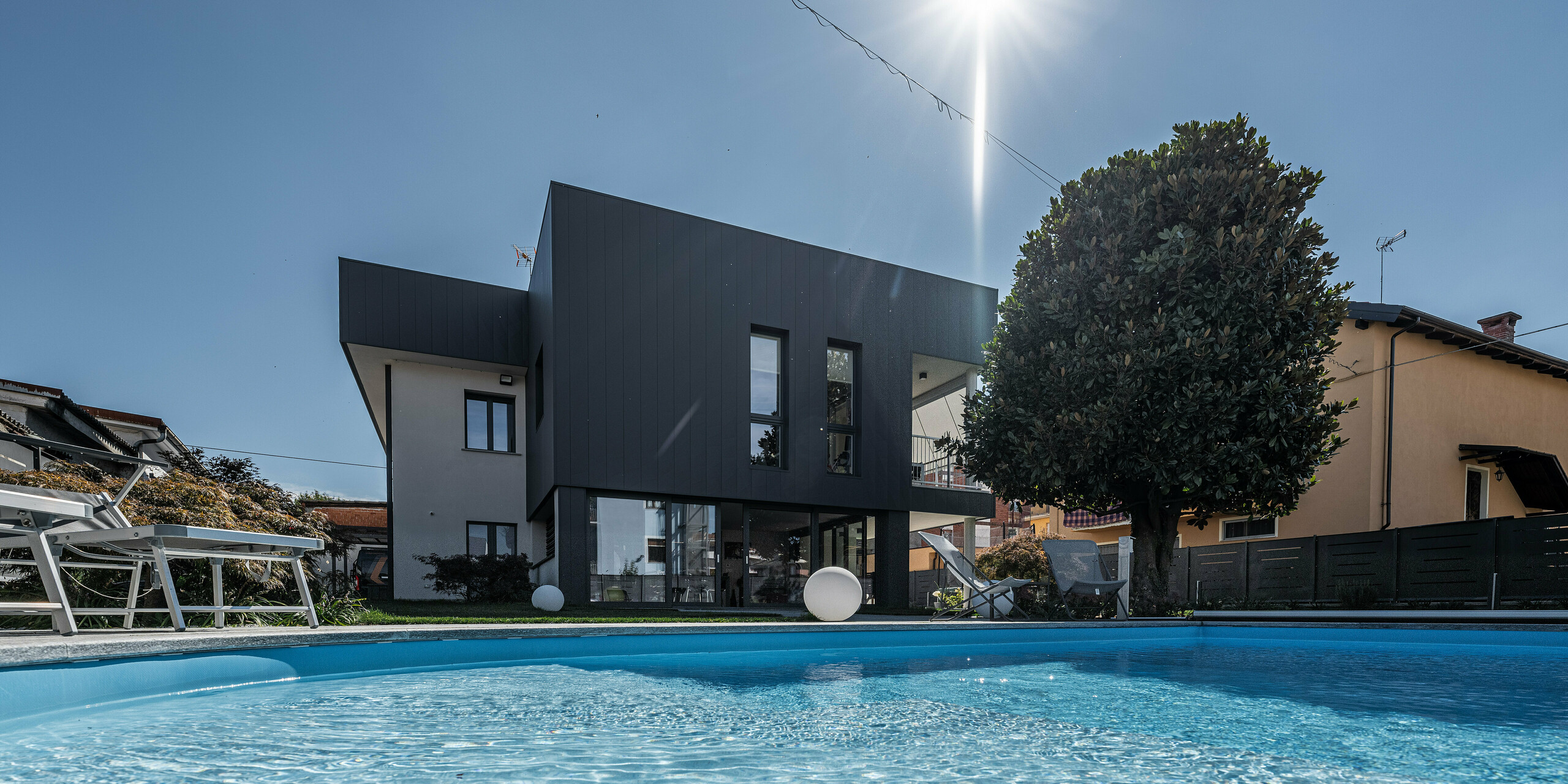 Eine ansprechende Ansicht eines Einfamilienhauses in Savigliano, Italien, vom blauen Wasser des Pools aus betrachtet, mit einer modernen PREFA Siding.X Aluminiumfassade in P.10 Dunkelgrau. Die Blechfassade präsentiert sich in klaren, scharfen Linien, die das zeitgenössische Design unterstreichen. Die Reflexion der Sonne im Wasser und die entspannende Poolumgebung betonen die luxuriöse und einladende Atmosphäre des Wohnortes.