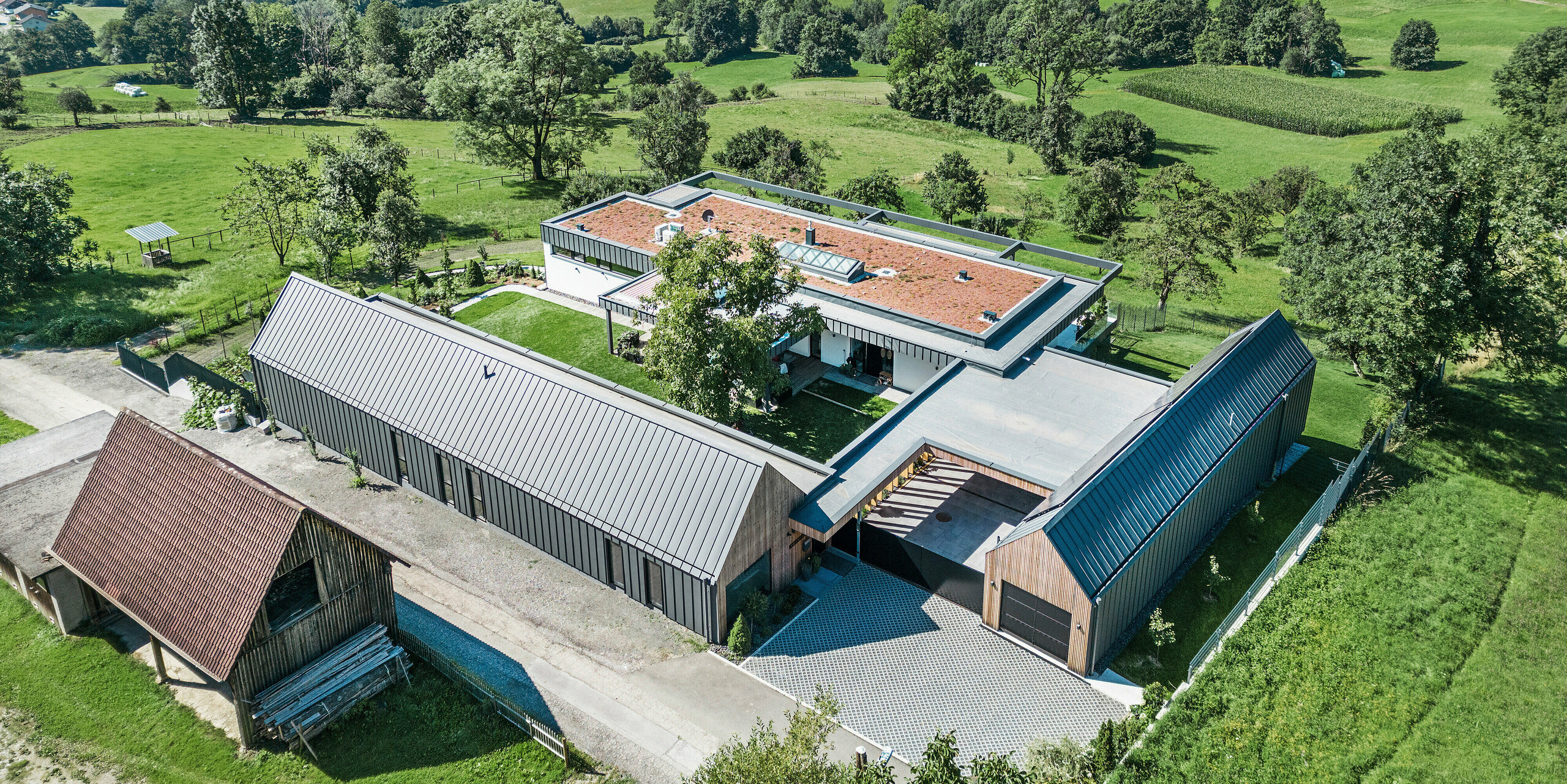 Luftaufnahme eines großzügigen Einfamilienhauses in Kombination mit landwirtschaftlichen Stallungen in Pogier, Österreich, eingebettet in eine malerische Landschaft mit Blick auf sanfte Hügel und grüne Felder. Die Gebäude sind mit einem PREFALZ Dach- und Fassadensystem in der Farbe dunkelgrau P.10 ausgestattet, das eine moderne Ästhetik in die ländliche Umgebung bringt. Die Anordnung der Gebäude schafft eine einladende Innenhofatmosphäre, während die Photovoltaikanlage auf einem der Dächer das Engagement des Hauses für Nachhaltigkeit und Energieeffizienz widerspiegelt.