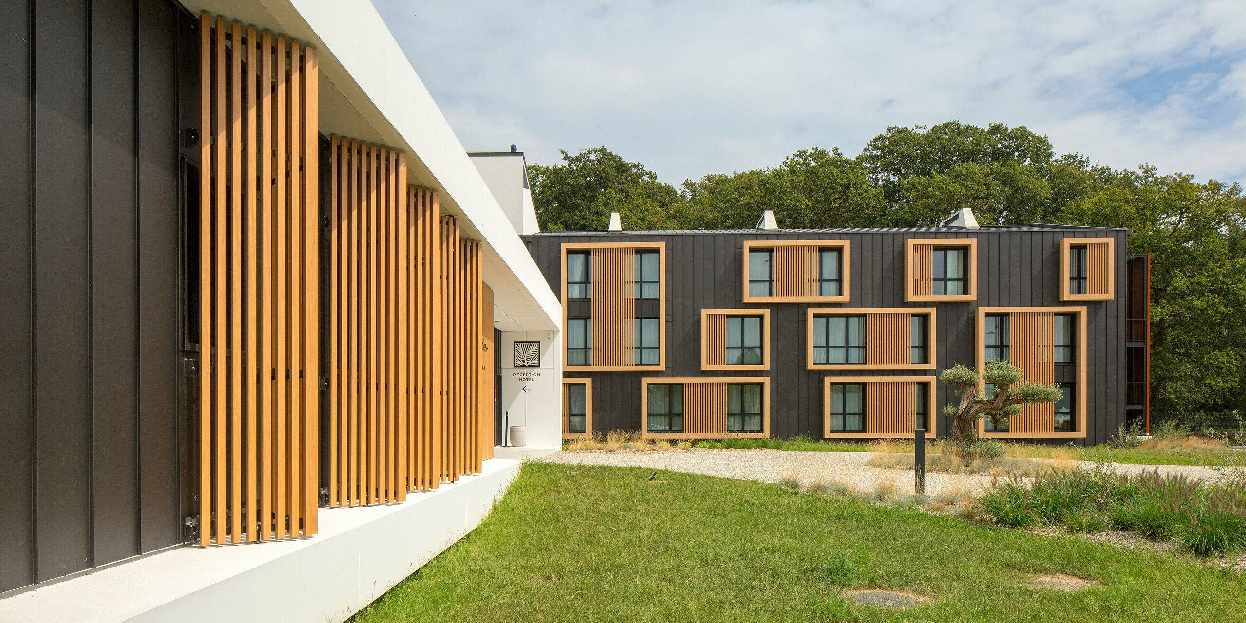 Moderne Fassade des Hôtel Parc du Landreau in Les Herbiers, Frankreich, mit einer Kombination aus Holz und Aluminium in Form von schwarzgrauem PREFALZ. Die markante Gestaltung mit Holzelementen und einer Stehfalzdeckung aus langlebigem, witterungsbeständigem Blech verleiht dem Gebäude einen zeitlosen architektonischen Charakter. Grünflächen und vertikale Fenster unterstützen das Gestaltungskonzept.