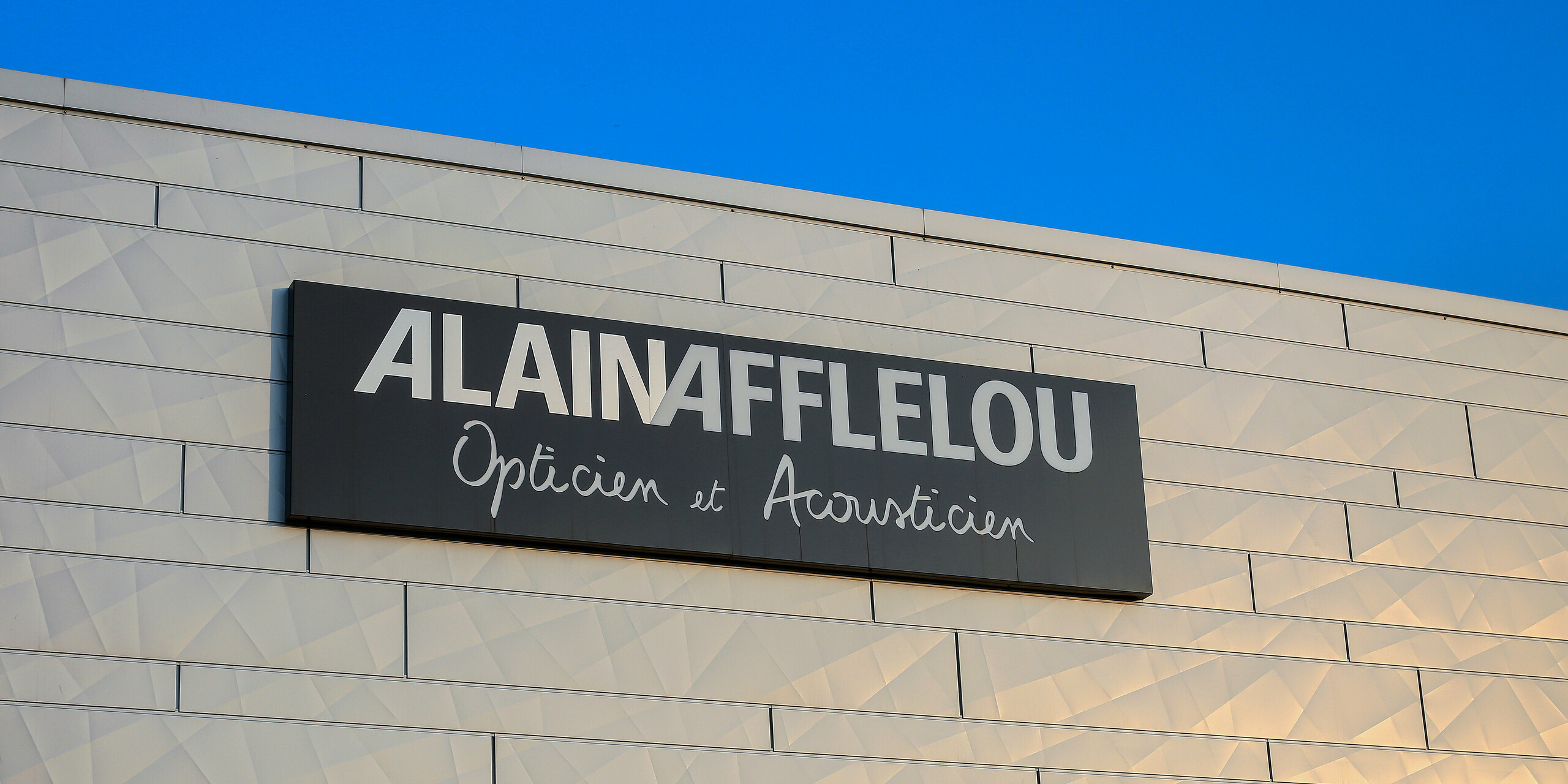 Gros plan sur l'enseigne de la société AFFLELOU sur le bâtiment de l'opticien à Auray - des panneaux de façade PREFA Siding.X argentés chatoyants sont visibles autour du logo.