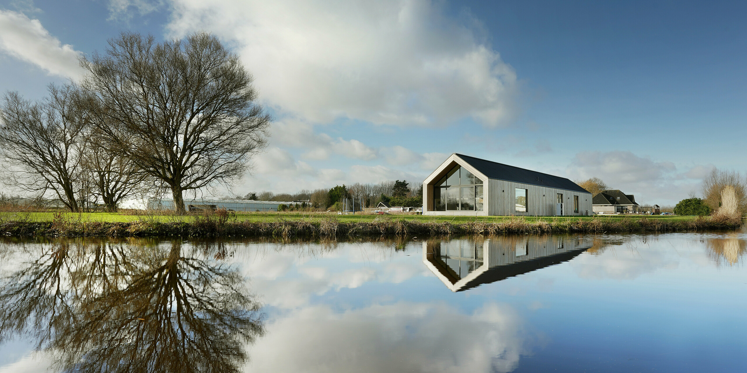 Panoramablick auf ein modernes Einfamilienhaus am Seeufer in Uithoorn, Niederlande, mit PREFALZ Winkelstehfalz-Eindeckung in Schwarzgrau. Das Bild zeigt das Gebäude mit großen Glasfronten und einer natürlichen Holz-Fassade, reflektiert im ruhigen Wasser des Sees. Die klare Linienführung und die nachhaltigen Materialien unterstreichen die durchdachte Bauweise. Ideal für innovative Bauprojekte, die auf ästhetisches Design und hohe Qualität setzen.