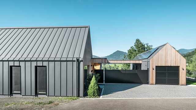 Eingangsbereich eines Einfamilienhauses mit angrenzender Garage in Pogier, Österreich, inmitten einer malerischen Landschaft. Dach und Fassade beider Gebäude sind mit langlebigem PREFALZ in P.10 dunkelgrau verkleidet, was ein modernes Erscheinungsbild erzeugt. Die Garage verfügt über eine harmonisch integrierte Holzverkleidung, die sich nahtlos in die umliegende Landschaft einfügt. Der gepflasterte Hof und die klaren Linien des Entwurfs ergänzen die Ästhetik des Grundstücks, während im Hintergrund die sanften Hügel das Bild einer friedlichen grünen Oase im grünen Herzen Österreichs vervollständigen.