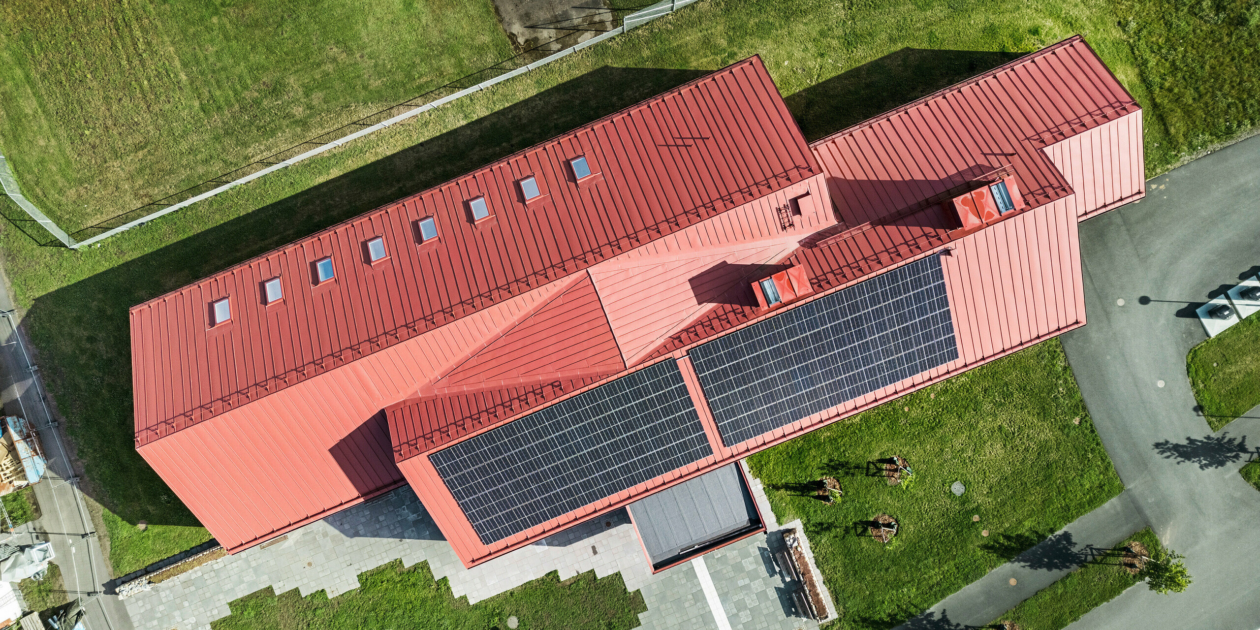 La vue aérienne de la cantine scolaire de Broby, en Suède, montre le toit frappant du bâtiment en PREFALZ en rouge oxyde. L'installation photovoltaïque à grande échelle et la toiture à joints debout clairement structurée en aluminium, qui confèrent au toit un aspect moderne et fonctionnel, sont clairement visibles. La disposition des fenêtres de toit laisse entrer beaucoup de lumière naturelle dans les espaces intérieurs. La vue plongeante offre une vue complète sur le bâtiment et une partie des espaces verts adjacents, qui soulignent l'intégration harmonieuse de la structure dans l'environnement.