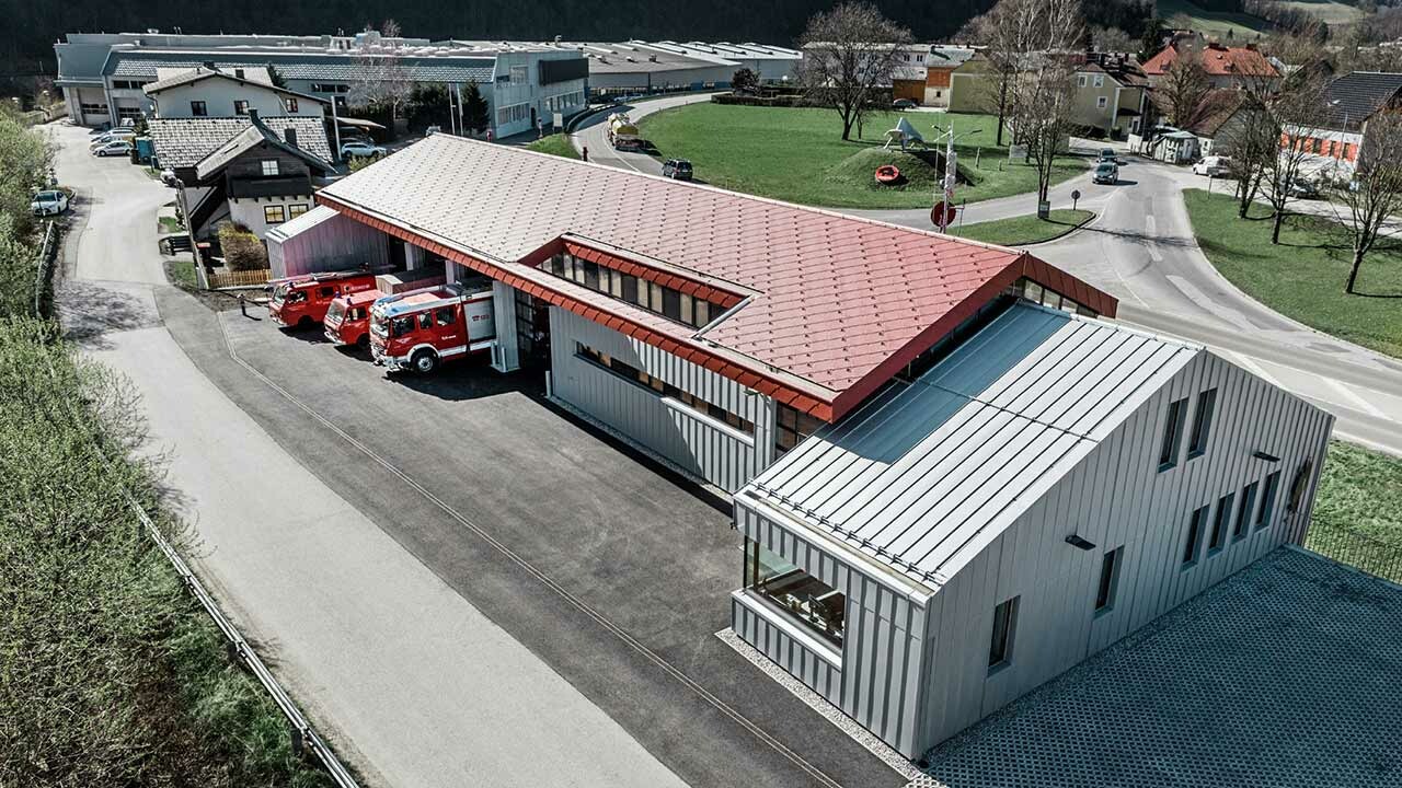 Vogelperspektive der Betriebsfeuerwehr Neuman und PREFA in Marktl, mit Feuerwehrautos in der Einfahrt, das Dach ist in Rot mit der großflächigen Aluraute eingedeckt, das Nebengebäude hat eine Gebäudehülle in Silbermetallic Blechbändern