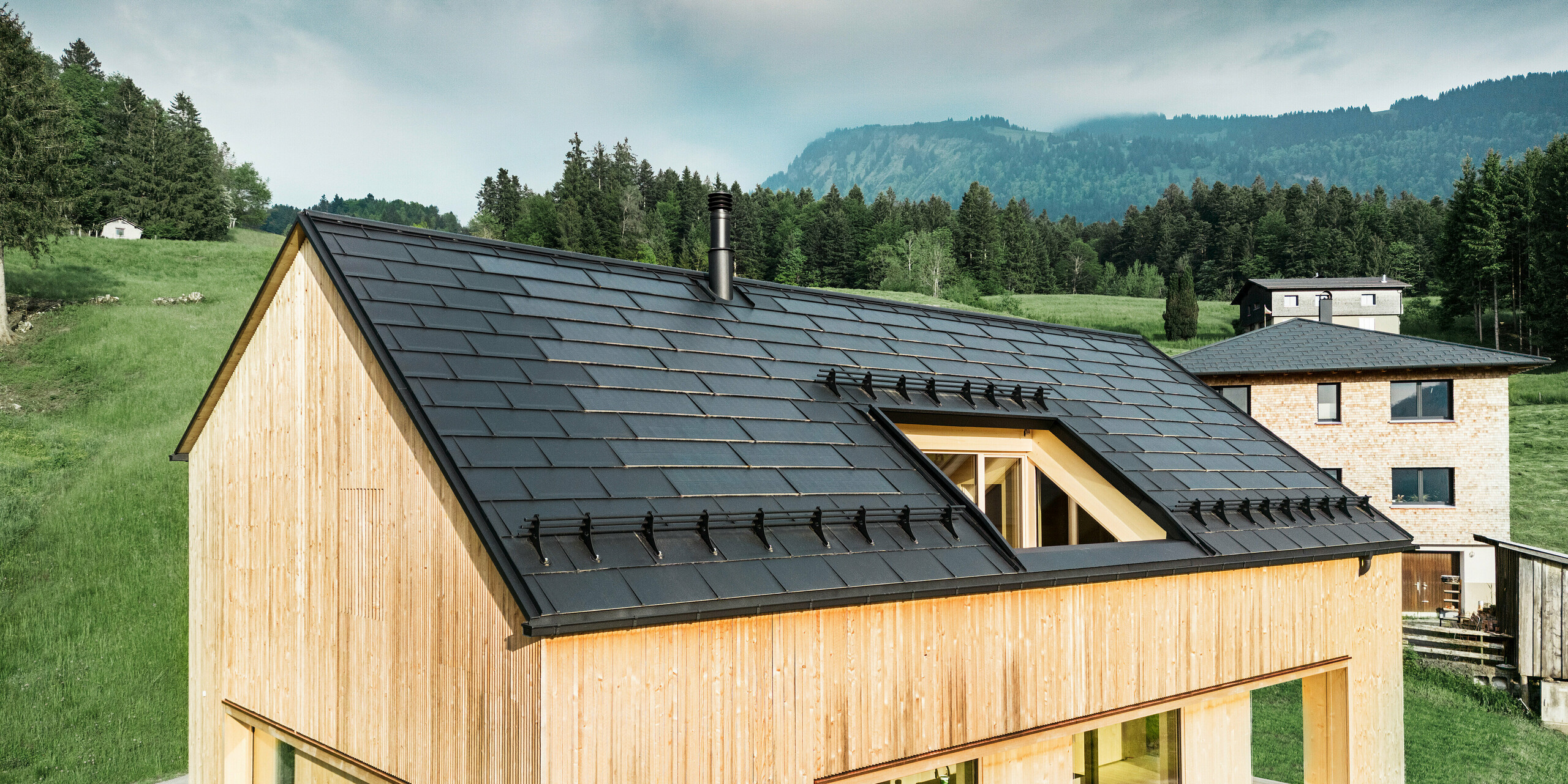 Vista frontale di una moderna casa in legno a Egg, che spicca sul paesaggio verde e rigoglioso con il suo tetto PREFA Solar scuro. Le tegole fotovoltaiche PREFA nere, posate con precisione, non solo forniscono protezione, ma anche efficienza energetica grazie alla tecnologia solare integrata. Le tradizionali tegole R.16 PREFA di colore nero sono state posate lungo i bordi del tetto fino alla gronda. Il camino classico e l'ampio lucernario completano il concetto di sostenibilità dell'edificio, che armonizza l'architettura moderna e le pratiche edilizie consapevoli dell'ambiente.