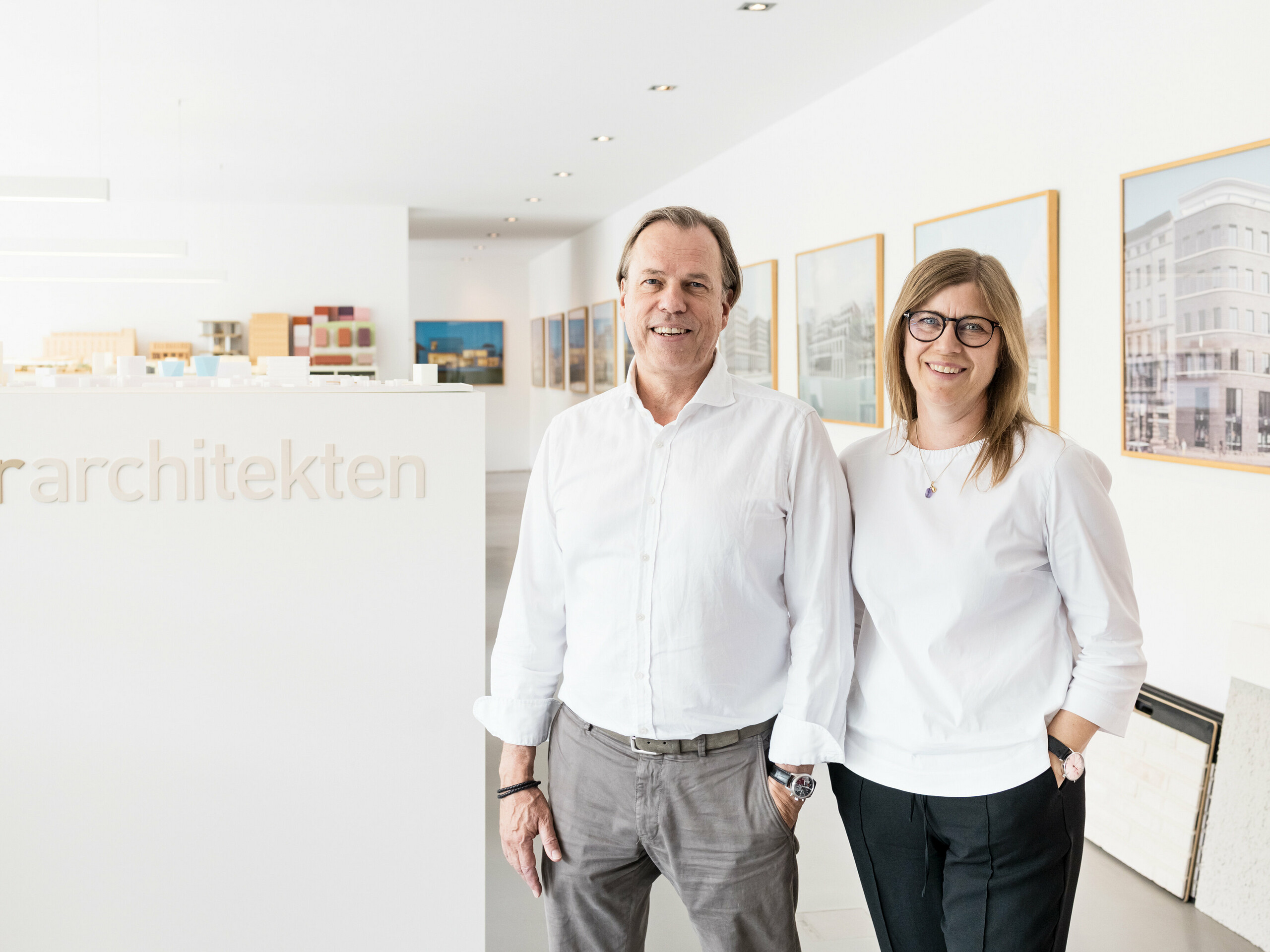 Leona e Andreas Geitner, gli architetti responsabili del progetto di Ratingen (DE), sono in piedi sorridenti davanti a una parete bianca con la scritta “Geitner Architekten”. Entrambi indossano camicie bianche, che conferiscono all'immagine un aspetto professionale e uniforme. Sullo sfondo sono visibili modelli architettonici e immagini incorniciate di progetti edilizi, che riflettono l'ambiente creativo e innovativo dello studio di architettura. L'atmosfera amichevole e invitante sottolinea la professionalità e la competenza del team di Leona e Andreas Geitner.