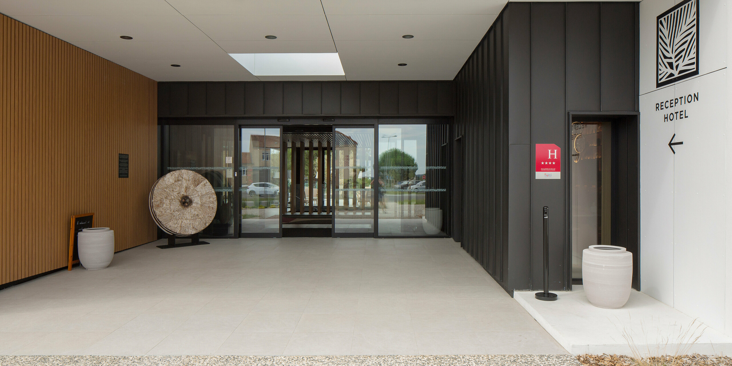 Modern gestalteter Eingangsbereich des Hôtel Parc du Landreau in Les Herbiers, Frankreich. Die Fassade ist mit schwarzgrauem PREFALZ verkleidet. Die Beschriftung am rechten Bildrand weist den Weg zur Hotelrezeption. Blumentöpfe, eine Holzverkleidung und die markante Stehfalzdeckung aus Aluminium verschönern den Hoteleingang, der über eine automatische Glasschiebetür zu erreichen ist. 