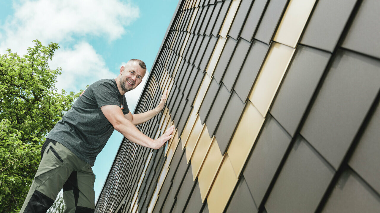 PREFA Handwerker mit grauem T-Shirt an einer PREFA Rauten-Fassade in Braun und Mayagold.