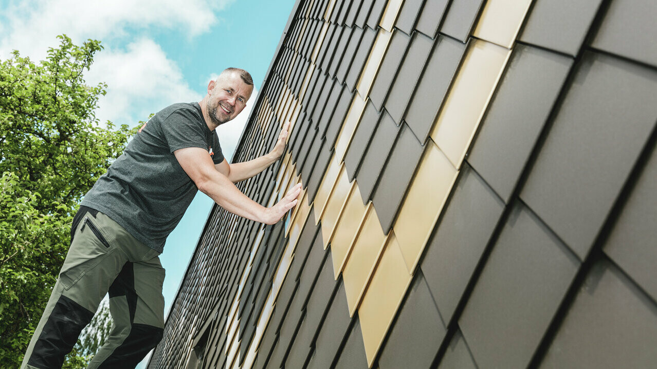 Artisan PREFA portant un t-shirt gris sur une façade en losanges PREFA couleurs brun et or maya.