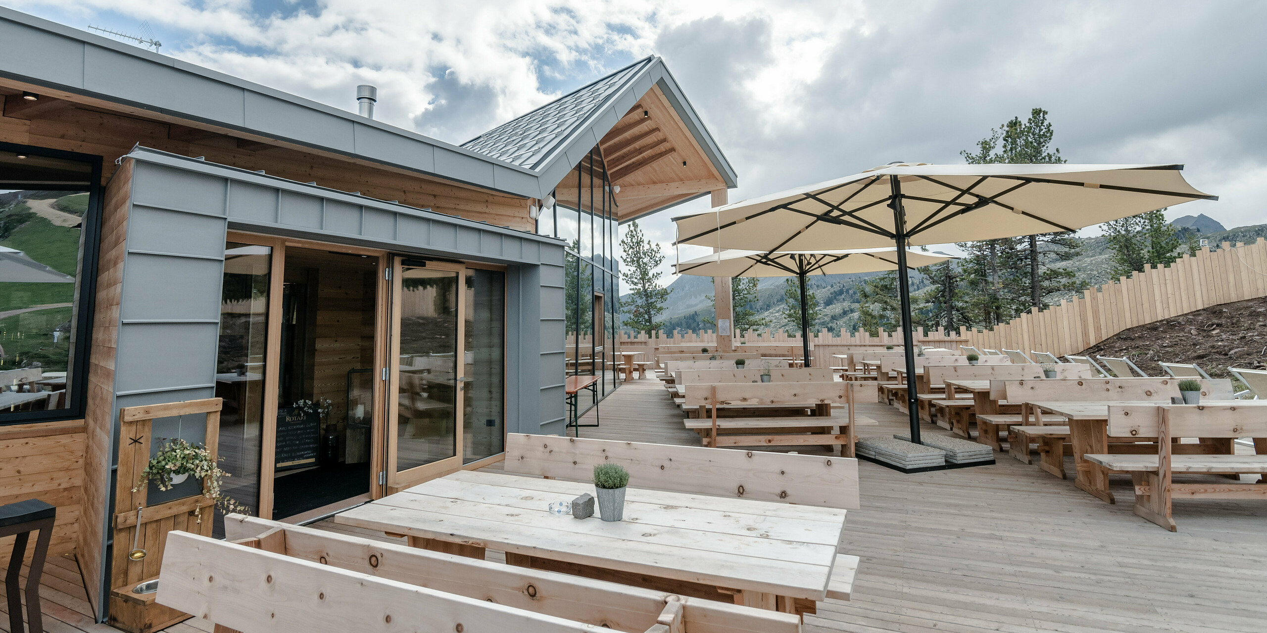 Das Ristorante Busabelle, umgeben von einer beeindruckenden Berglandschaft, präsentiert eine weitläufige Terrasse mit robusten Holztischen und eleganten Sonnenschirmen, die für gemütliche Sitzmöglichkeiten sorgen. Die Architektur kombiniert eine moderne, hellgraue PREFALZ Fassade mit natürlichen Holzelementen und einem spitz zulaufenden Dach, was dem Gebäude eine frische und einladende Atmosphäre verleiht.