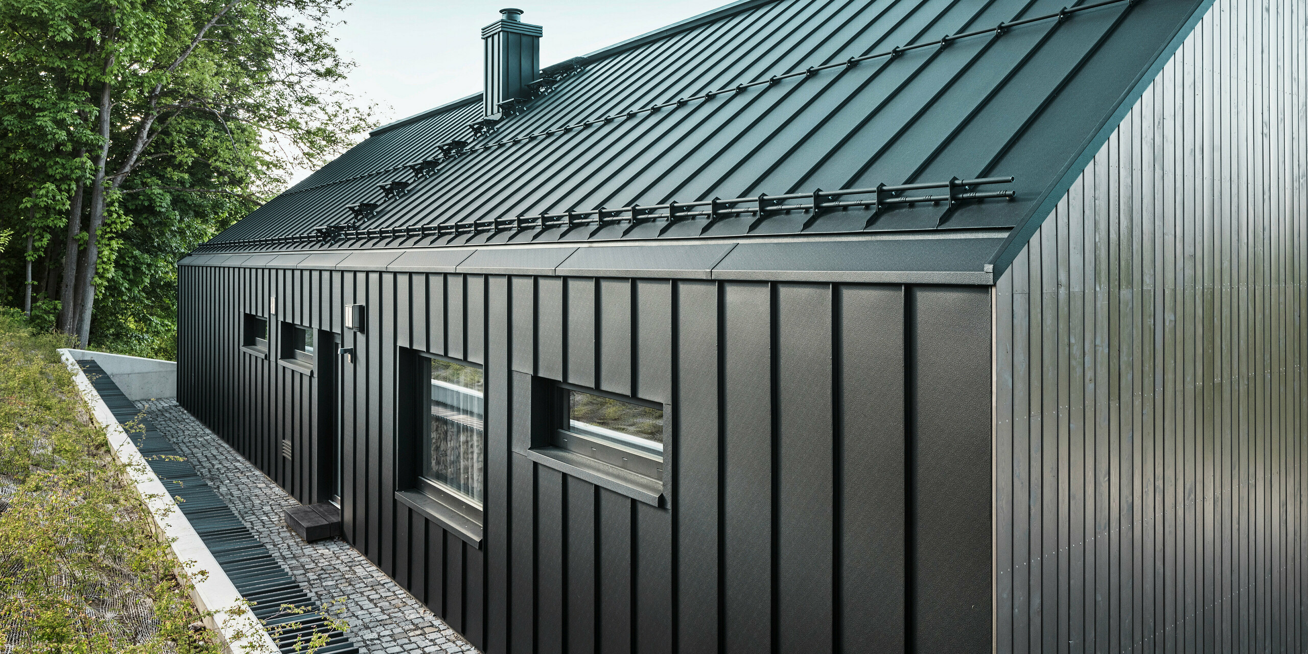 Nahaufnahme der Rückseite eines modernen Einfamilienhauses in Kuncice, CZ. Das dunkle Holzmassivhaus wurde am Dach und der Gebäuderückseite großflächig mit PREFALZ in P.10 Schwarz verkleidet. Auf der Rückseite sind drei horizontale sowie ein quadratisches Fenster angebracht worden. Zwischen den vier Fenstern ist eine Tür mit einer kleinen Stufe zu sehen.