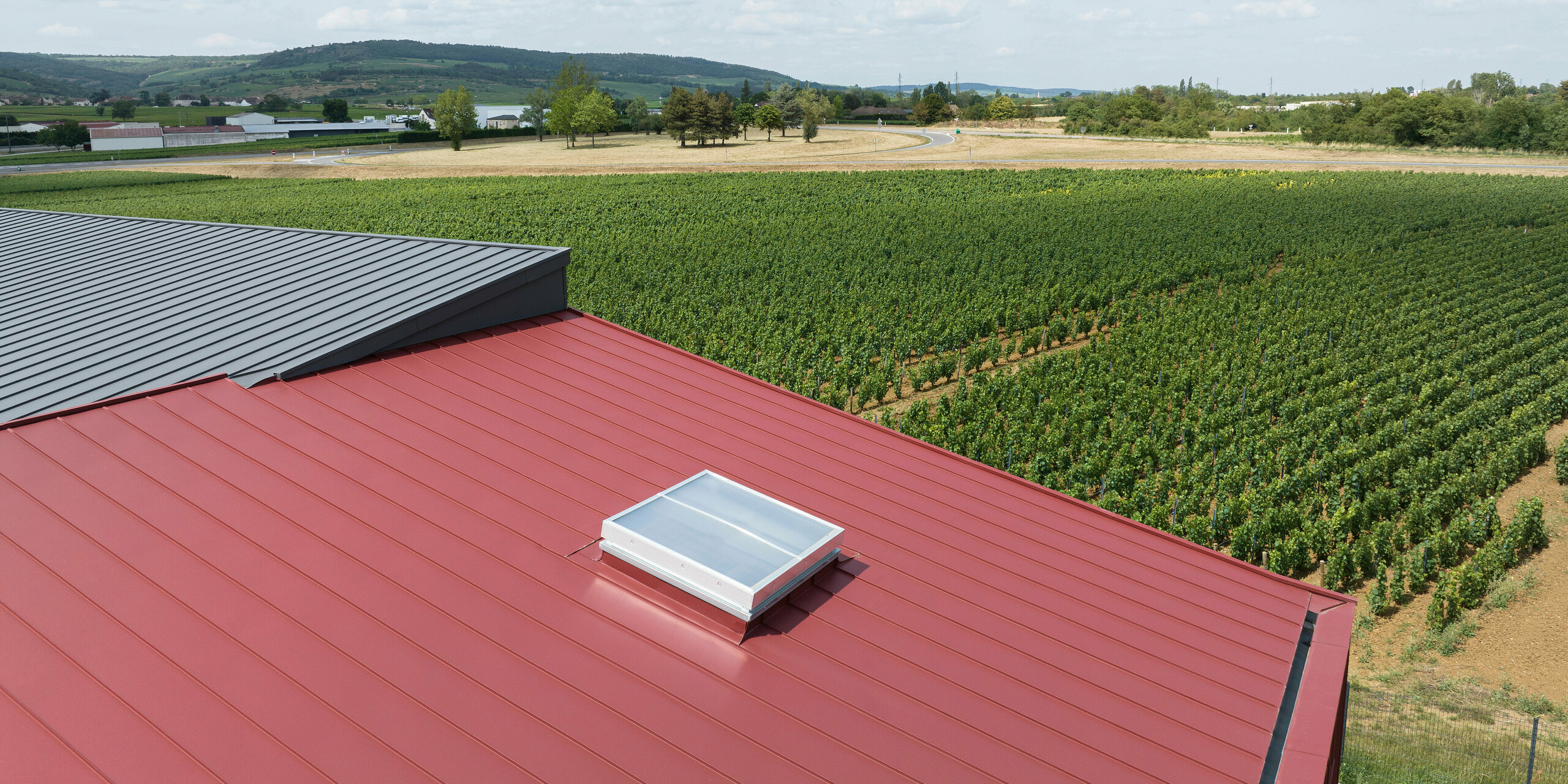 L'image montre une scène rurale avec un toit en aluminium de deux couleurs différentes. Une lucarne est installée sur la partie rouge oxyde du toit, indiquant une structure spatiale habitable en dessous. La zone gris sombre du toit est plutôt petite sur le bord gauche de l’image. En arrière-plan, vous pouvez voir les zones agricoles d'un domaine viticole. La composition de l'image vise à montrer l'intégration des systèmes modernes de toiture en aluminium dans les environnements ruraux.