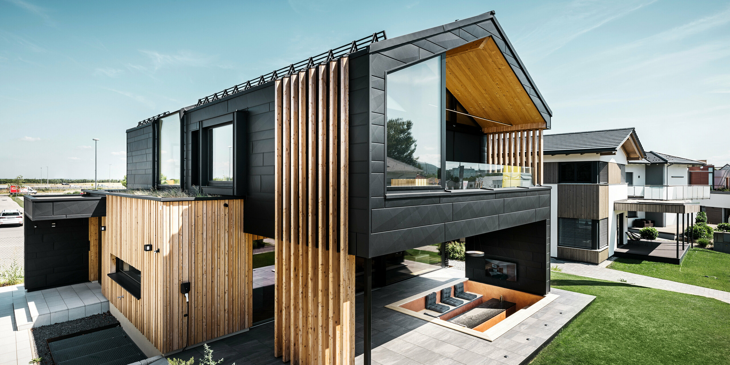 Vue latérale de la maison modèle Autark dans le Blue Lagoon à Vösendorf, en Autriche. L'étage supérieur impressionne par ses grandes surfaces vitrées et sa façade moderne en aluminium composée de panneaux PREFA Siding.X en P.10 noir, associés à des éléments verticaux en bois. La terrasse spacieuse au rez-de-chaussée avec sièges encastrés s'intègre parfaitement dans l'architecture. Le design élégant et durable de la maison combine de manière impressionnante l'aluminium et le bois.