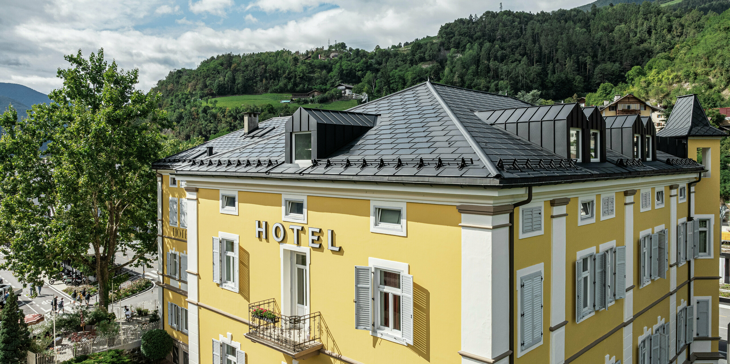 Drohnenaufnahme des Hotels Jarolim in Brixen mit einem langlebigen Aluminiumdach aus PREFA Solardachplatten und Dachplatten R.16 in eleganter schwarzer Optik mit hochwertiger P.10 Beschichtung. Das robuste und witterungsbeständige PREFA Dach vereint Ästhetik und Funktionalität und ist ideal für denkmalgeschützte Gebäude. Die harmonische Integration von Photovoltaik-Modulen in die robusten Aluminium-Dachplatten ermöglicht eine nachhaltige Energiegewinnung, ohne die historische Architektur zu beeinträchtigen. Die präzise Verarbeitung unterstreicht den hochwertigen Charakter der PREFA Produkte.