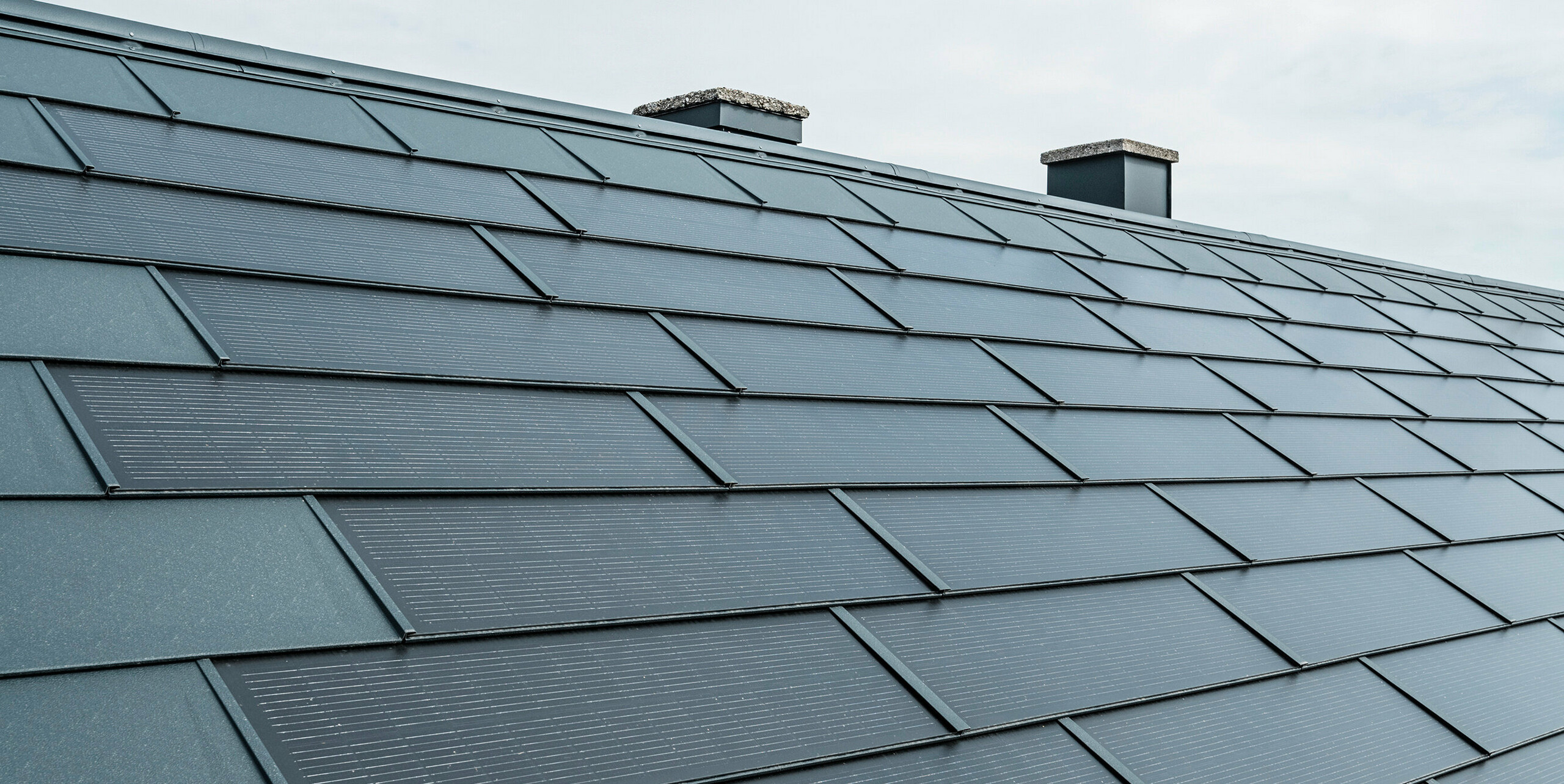 Vue en perspective d'une toiture équipée de tuiles solaires PREFA de couleur anthracite. Les panneaux sont posés avec précision et offrent une solution innovante de production d'énergie grâce à leurs cellules photovoltaïques intégrées. Deux cheminées s'élèvent à l'arrière-plan, surplombées par un ciel nuageux, soulignant la combinaison de l'architecture de toiture traditionnelle et de la technologie solaire moderne.