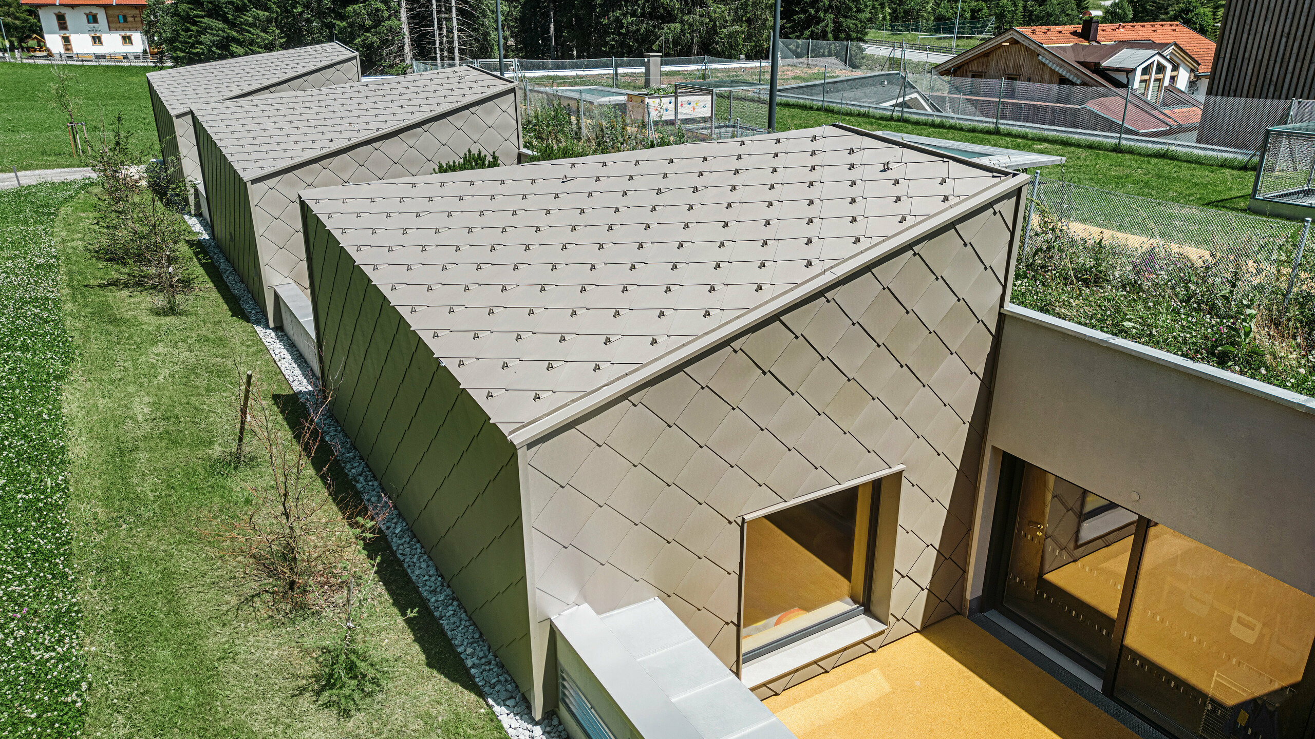 Des losanges de toiture et de mur PREFA 44 × 44 de couleur bronze couvrent le toit et une partie de la façade d'un bâtiment moderne à Gerlos, dans la vallée du Ziller. Le bâtiment dispose de grandes baies vitrées et de zones de terrasse. En arrière-plan, on peut voir l'environnement rural avec des prairies verdoyantes et une colline boisée.