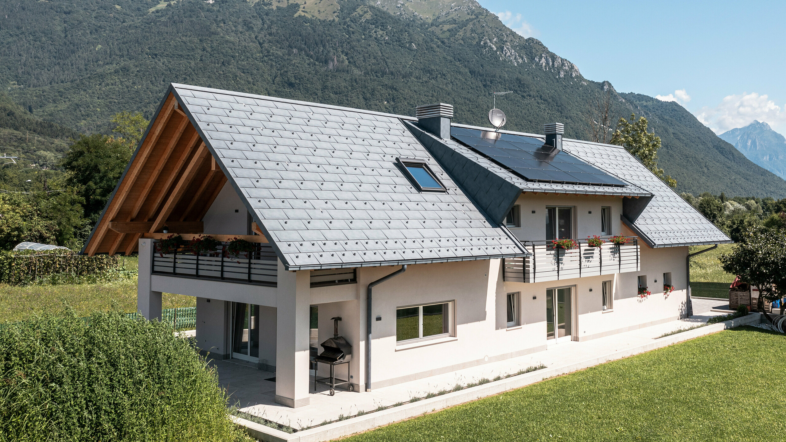 Drohnenaufnahme eines Hauses in Belluno, Italien, eingedeckt mit PREFA R.16 Dachplatten in P.10 anthrazit. Das Anwesen liegt in einer atemberaubenden Alpenlandschaft und kombiniert traditionelle Elemente wie freiliegende Holzgiebel mit modernen, langlebigen Baumaterialien, wodurch eine perfekte Symbiose aus Tradition und Moderne entsteht.