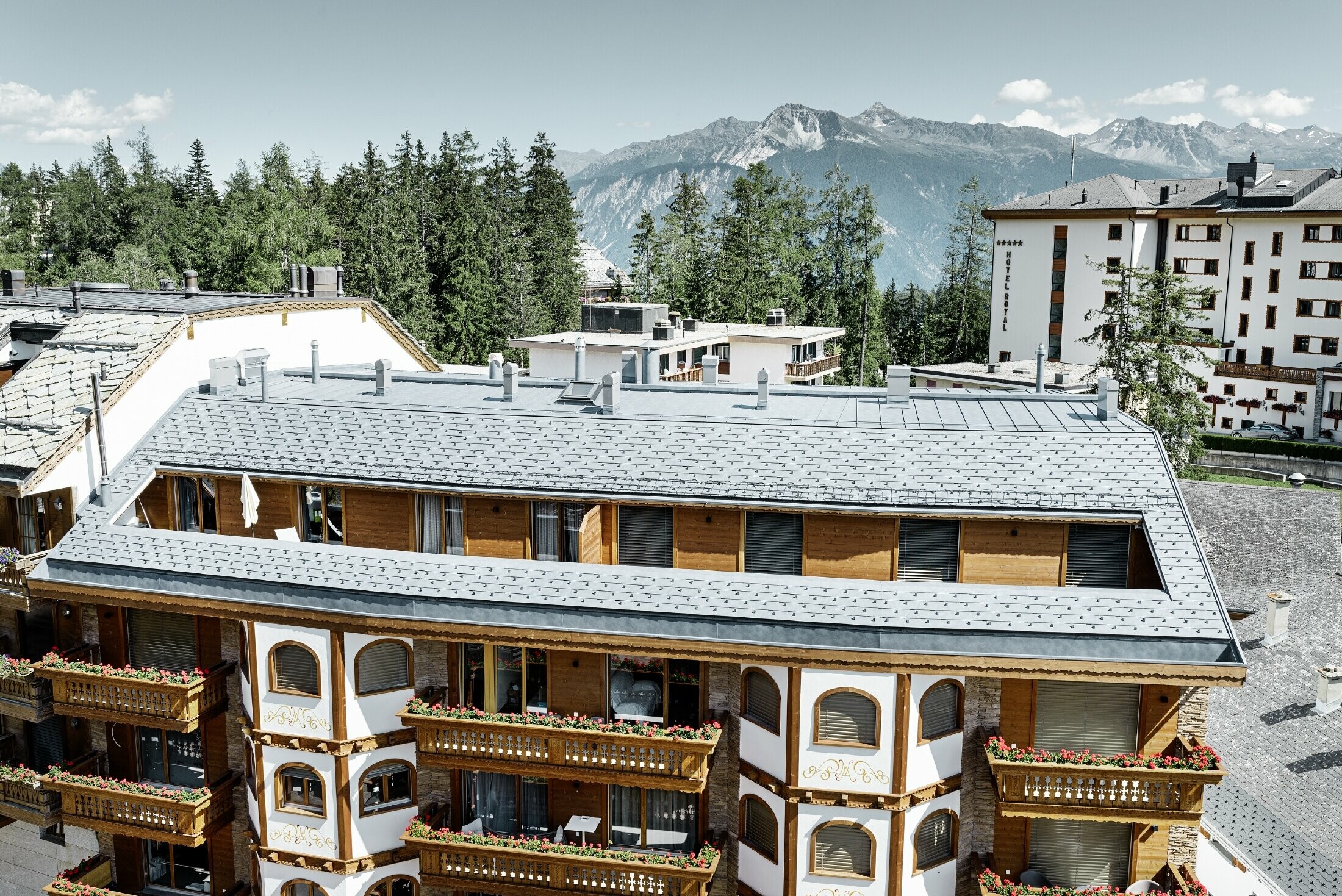 Casa plurifamiliare in Crans-Montana con montagne sullo sfondo e una facciata con elementi decorativi in legno e con una copertura in scandole PREFA grigio pietra