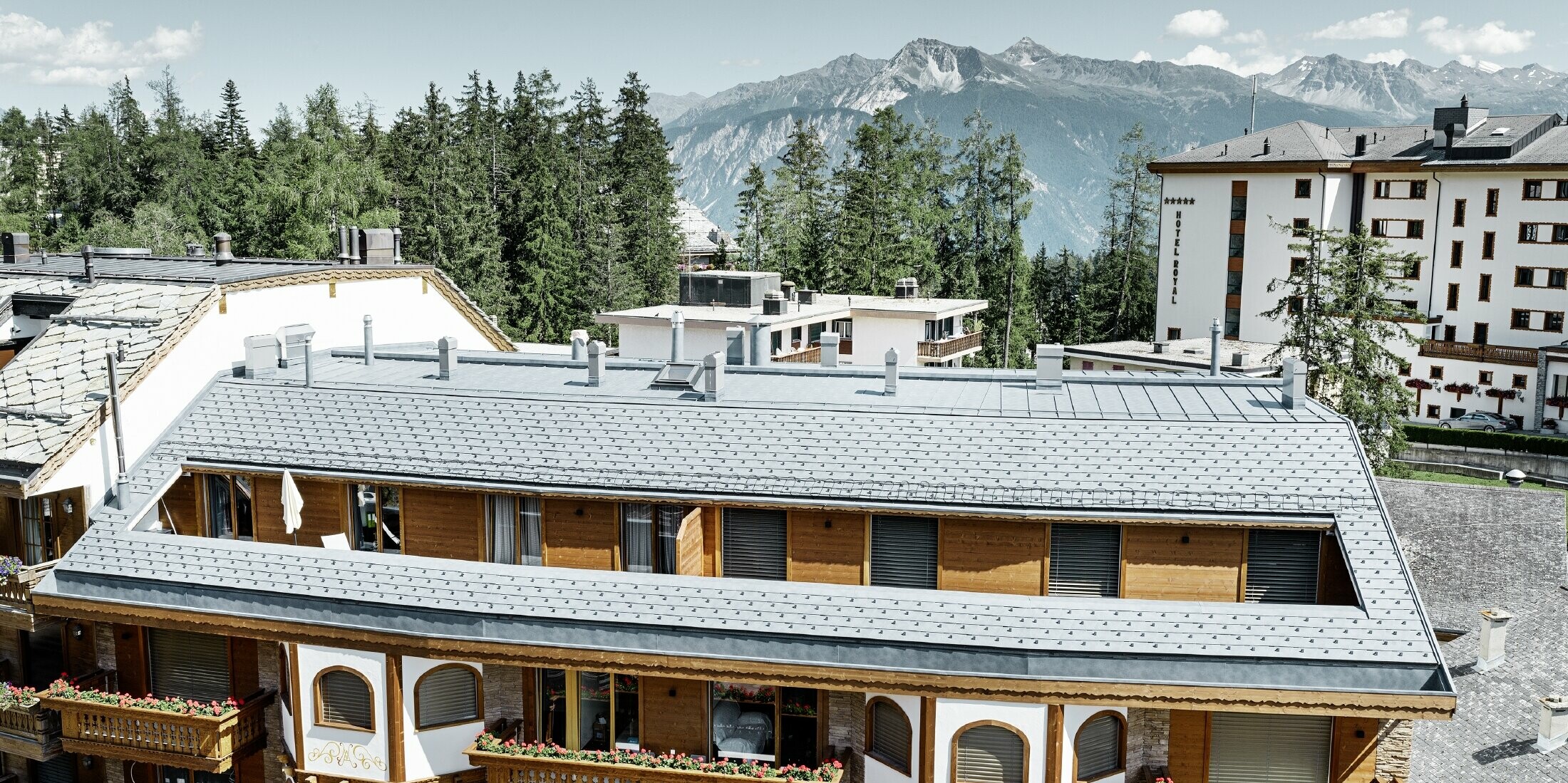 Mehrfamilienhaus in Crans-Montana mit Bergen im Hintergrund und einer Fassade mit verspielten Holzelementen mit einem PREFA Aluminium Dachschindel-Dach in steingrau 