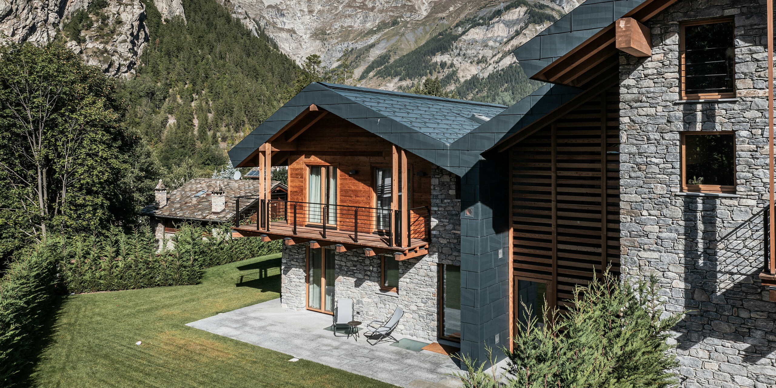 Prächtige Villa in Courmayeur, eingebettet in die atemberaubende Berglandschaft des Aostatals. Die Fassade kombiniert elegant Holz, Stein und PREFA Aluminium in Steingrau. Die Villa zeichnet sich durch mehrere Holzbalkone und eine großzügige Terrasse aus, die zum entspannten Genuss der spektakulären Aussicht auf die umgebenden Gipfel einlädt. Üppiges Grün und die historischen Steinhäuser im Hintergrund vervollständigen das idyllische Alpenpanorama. Die robusten und langlebigen Dach- und Fassadenpaneele FX.12 aus PREFA Blech bieten dem Gebäude starken Schutz vor winterlichen Bedingungen.