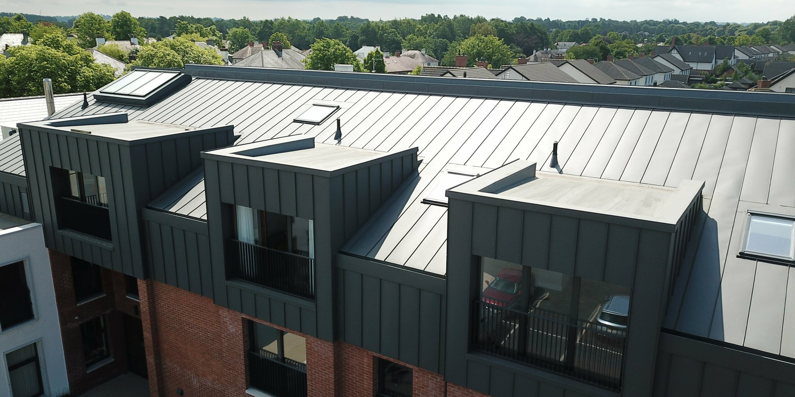 Vogelperspektive auf das Dach der Harberton Hall in Belfast, Nordirland. Das moderne Gebäude für betreutes Wohnen kombiniert PREFALZ Aluminium in P.10 Dunkelgrau mit einer klassischen Ziegelfassade. Die Flachdachgauben mit ihren großen Fenstern sorgen für lichtdurchflutete Innenräume. Die Stehfalzverkleidung schafft eine harmonische Verbindung von zeitgemäßem Design und traditioneller Bauweise.