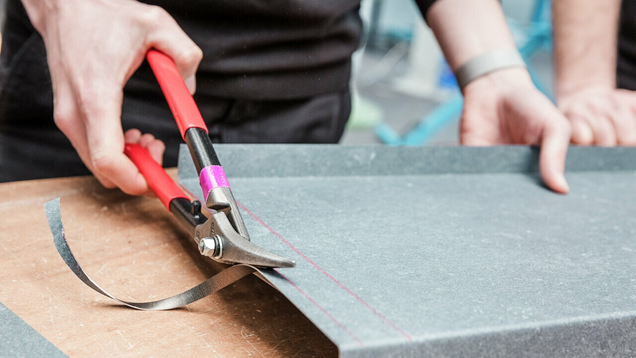 Ein Teilnehmer der PREFA Academy schneidet mit einer Blechschere präzise eine Aluminiumplatte entlang einer markierten Linie. Die Szene zeigt praktische Schulungsmaßnahmen, bei denen handwerkliche Fähigkeiten im Umgang mit Aluminium vermittelt werden.