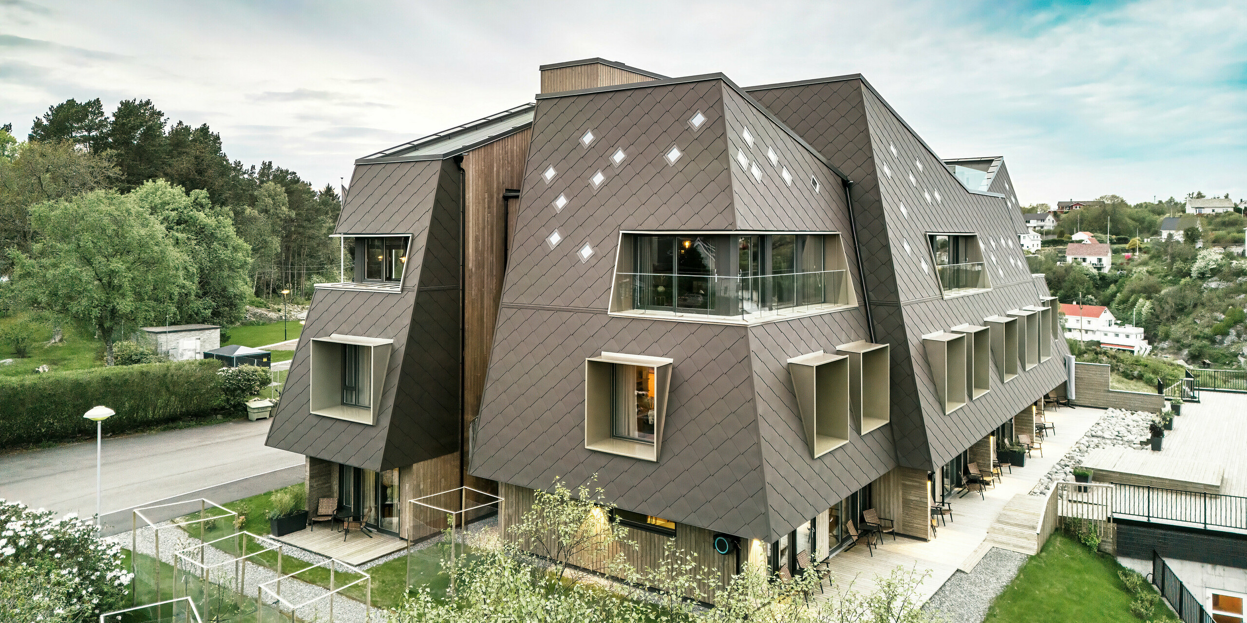 Stirnseite des Beckerwyc House in Bekkjarvik, Norwegen, mit einer markanten Fassade aus PREFA Wandrauten 44 × 44 in Nussbraun. Das Bild zeigt die futuristische Architektur des Gebäudes mit schrägen Dachflächen und hervorspringenden Fensterelementen. Die großzügigen Terrassen und Balkone sowie die umgebende grüne Landschaft betonen das harmonische Zusammenspiel von Natur und moderner Bauweise. Die robuste Gebäudehülle aus hochwertigem Aluminium sorgt für langlebigen Schutz.