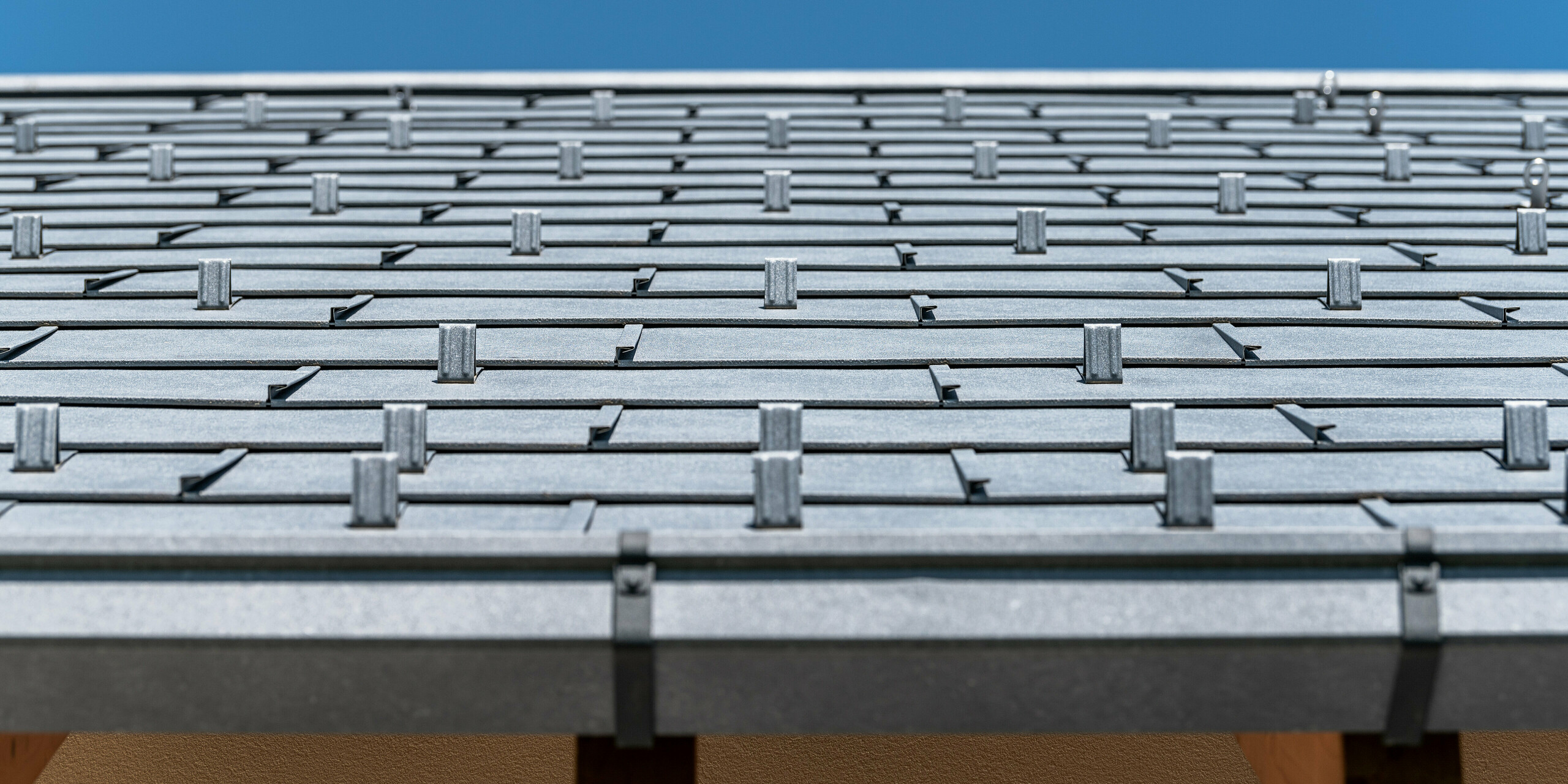 Detailansicht des PREFA Daches eingedeckt mit Dachpaneel R.16 in Farbe P.10 anthrazit. Das Bild zeigt präzise verarbeitete Aluminium-Dachplatten. Die Aufnahme hebt die markante Struktur und das moderne Design des Daches hervor, das im Kontrast zum strahlend blauen Himmel steht und die innovative und langlebige Dachlösung von PREFA unterstreicht.