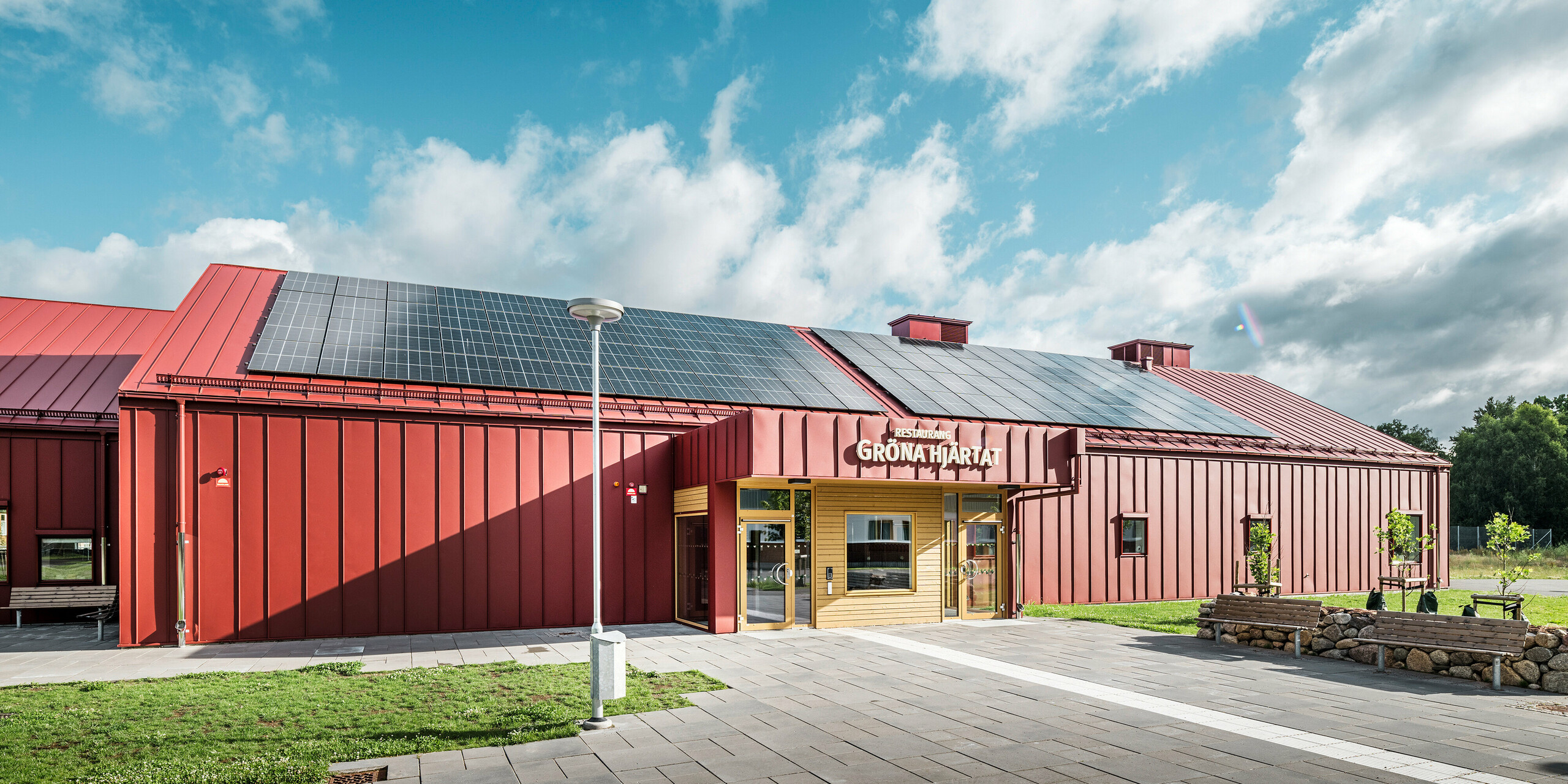 Vue de la zone d'entrée du restaurant Gröna Hjärtat, cantine scolaire de Göingeskolan et Prästavångskolan à Broby, Suède. Le bâtiment a reçu une façade remarquable en PREFALZ en rouge oxyde. Un système photovoltaïque à grande échelle fournissant de l'énergie renouvelable a été installé sur le toit. La combinaison d'un revêtement en aluminium durable et d'un design sophistiqué fait de ce bâtiment un exemple exceptionnel d'architecture moderne. La gouttière PREFA est également conçu en rouge oxyde, semblable au toit et à la façade, et souligne l'aspect harmonieux du bâtiment.