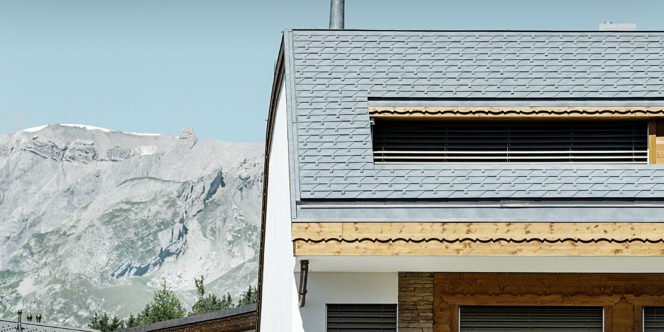 Mehrfamilienhaus in Crans-Montana mit Bergen im Hintergrund und einer Fassade mit verspielten Holzelementen mit einem PREFA Aluminium Dachschindel-Dach in steingrau 