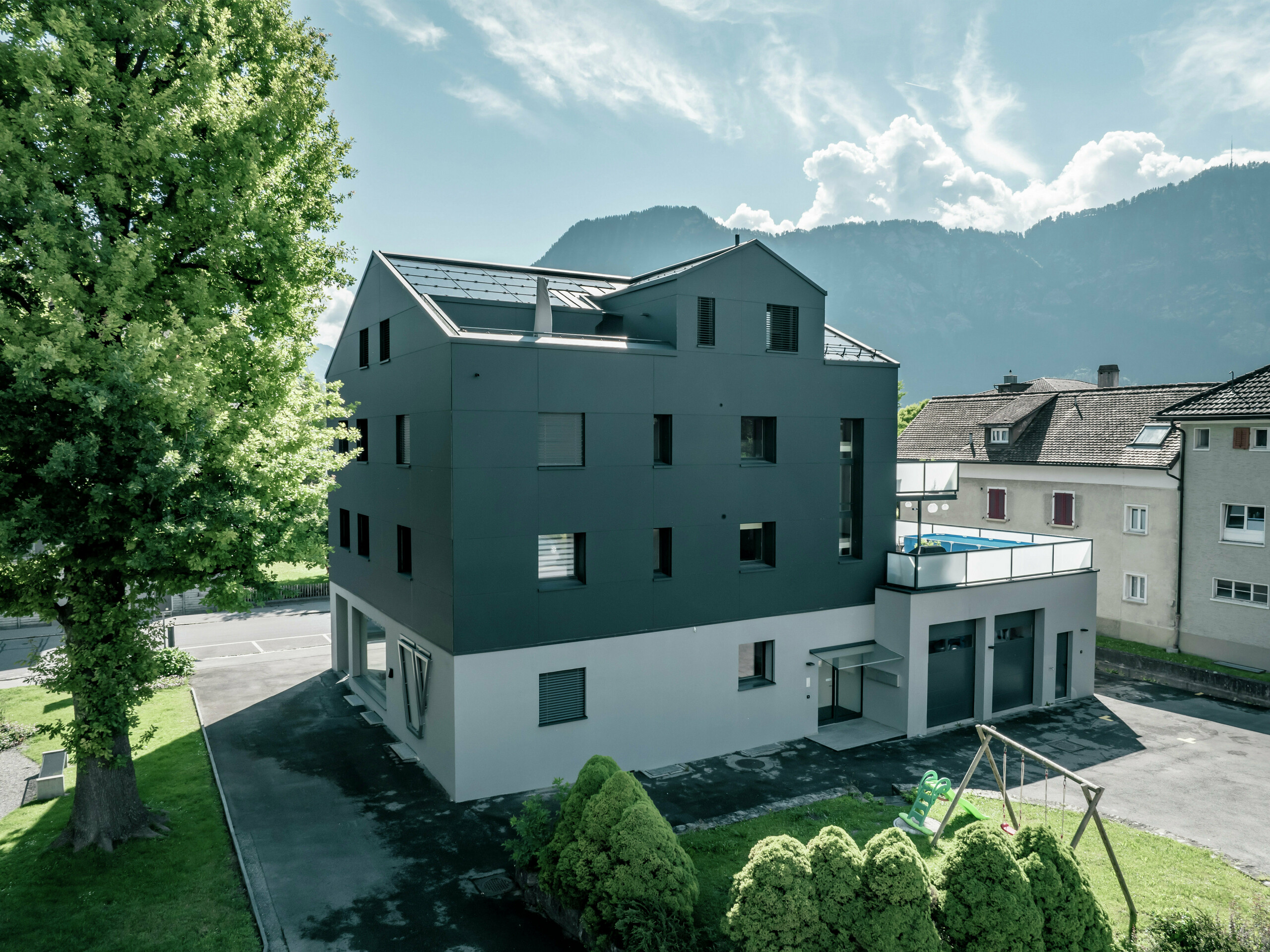 Modernisiertes Wohn- und Geschäftsgebäude mit einer anthrazitfarbenen PREFA Fassade aus PREFABOND Aluminium Verbundplatten. Die Seitenansicht zeigt klare Linien und ein zeitgemäßes Design. Im Vordergrund befinden sich gepflegte Grünflächen und ein kleiner Spielplatz. Im Hintergrund sind Berge und ein sonniger Himmel mit leichten Wolken erkennbar.