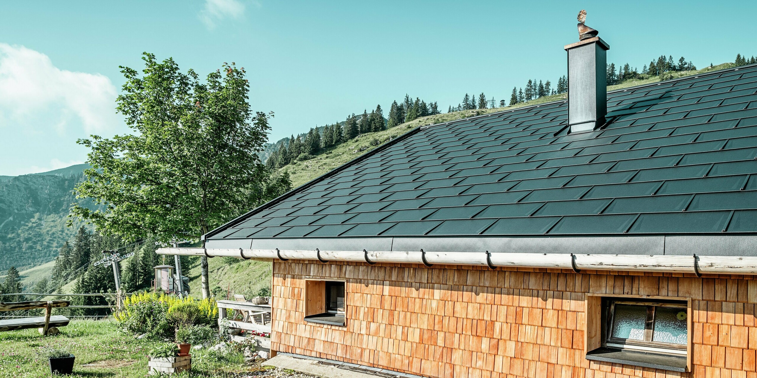 Vue latérale de la Birkehütte sur le Walleralm, Bayrischzell, après la rénovation complète du toit avec des panneaux de toit PREFA durables R.16 en P.10 noir et des tuiles solaires PREFA innovantes. Le charme classique de la façade en bois est complété par le systèmes d'évacuation des eaux intéressant du toit avec une combinaison de gouttière en bois et de tuyau de descente en aluminium. L'élégante cheminée est revêtue d'aluminium et est ornée d'une figure en métal au point le plus élevé. Cette figure artistique symbolise un chapeau de chasse traditionnel. La figure artistique symbolise un chapeau de chasse traditionnel. La combinaison harmonieuse de la technologie moderne et de l'architecture traditionnelle s'intègre parfaitement dans le paysage montagneux pittoresque.