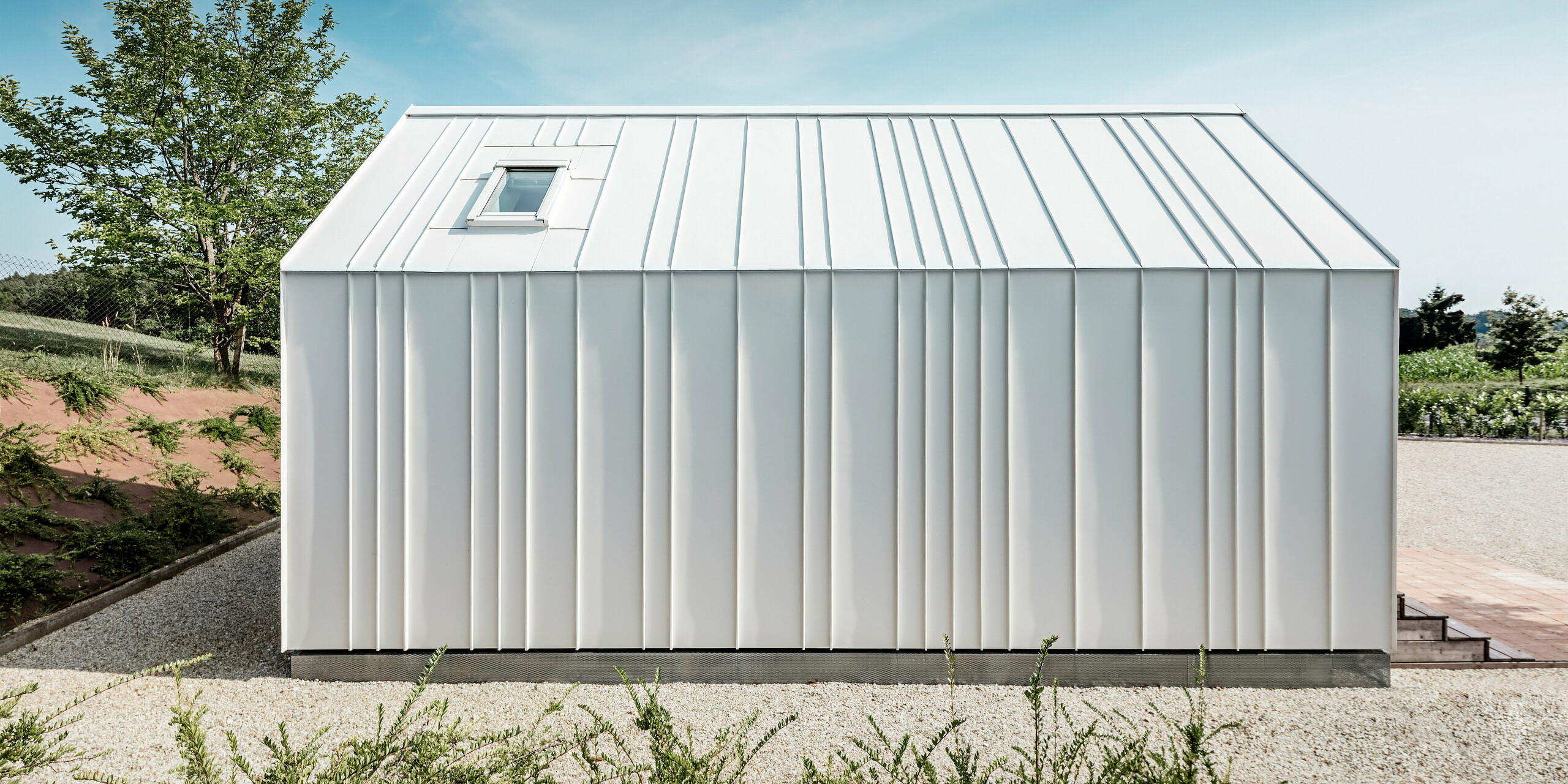 Vue latérale de l'une des petites maisons « Pri Momi » à Kančevci, en Slovénie. La micro-maison est recouverte d'un élégant toit à joints debout fabriqué à partir du système de toiture et de façade durable PREFALZ. Le revêtement en aluminium de haute qualité est de couleur blanc pur P.10. L'image montre les plis exécutés avec précision et les lignes élégantes de la coque en aluminium blanc, qui confèrent au bâtiment un aspect moderne et minimaliste. L'environnement naturel et le design clair et simple s'harmonisent parfaitement.