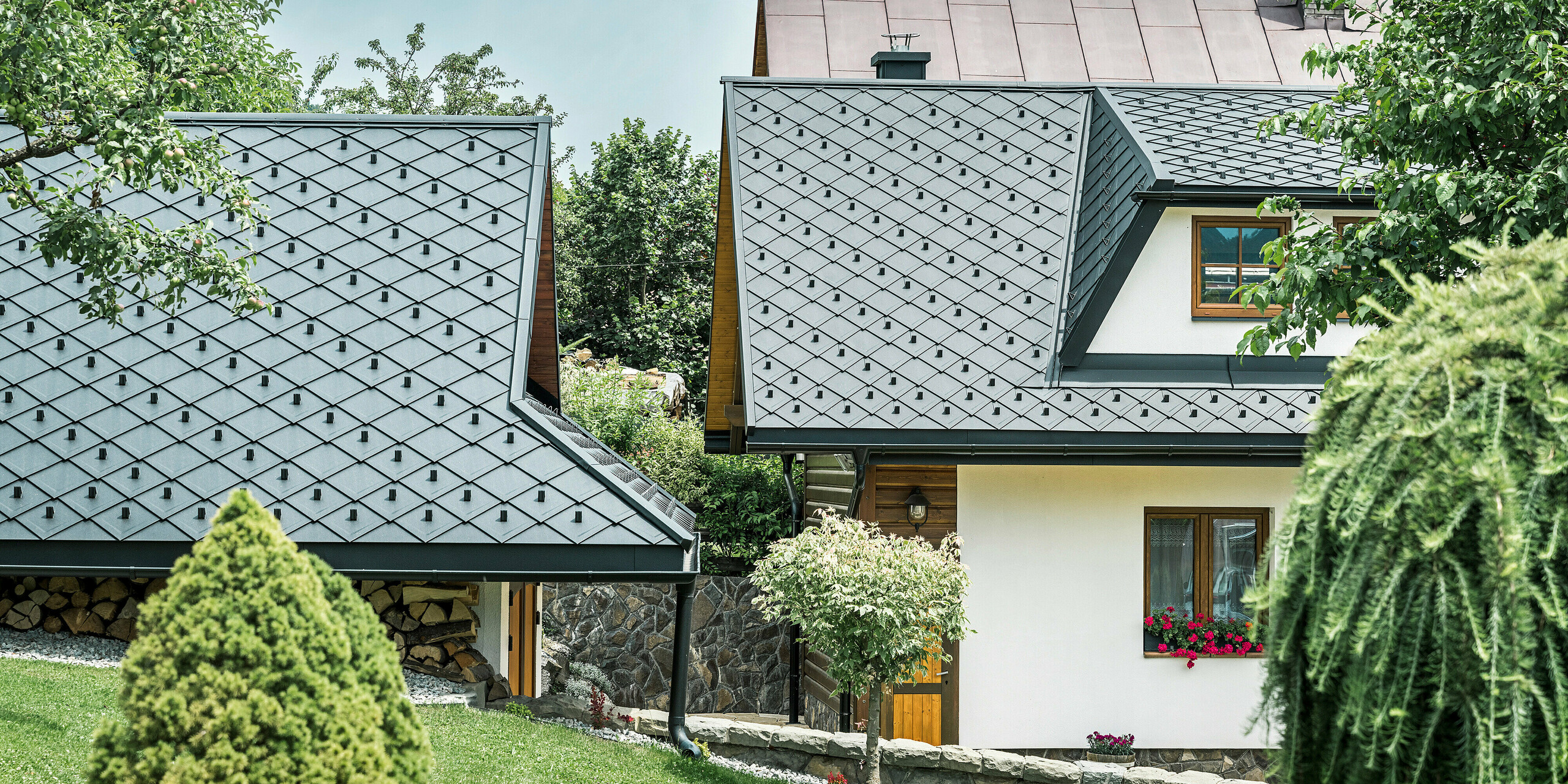 Vue détaillée des toits d'une maison familiale et d'une extension à Stara Bystrica, Slovaquie, recouverts d'une toiture en losanges PREFA 29 × 29 en P.10 noir. L'aménagement du toit présente un aspect élégant, obtenu grâce à des losanges en aluminium robustes. Les lignes claires et le design harmonieux des toits s'intègrent parfaitement dans l'environnement et soulignent l'aspect moderne et élégant du bâtiment.