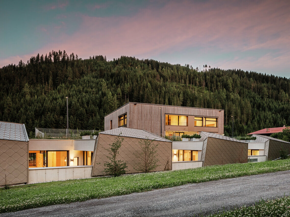 La photo montre le centre éducatif de Gerlos à la tombée de la nuit, avec l'éclairage intérieur. Avec ses boiseries modernes et ses panneaux métalliques en forme de losange, le bâtiment s'intègre harmonieusement dans le cadre naturel de la forêt dense de conifères à l'arrière-plan. Le ciel baigne dans les tons doux du soir, créant une atmosphère calme et invitante.