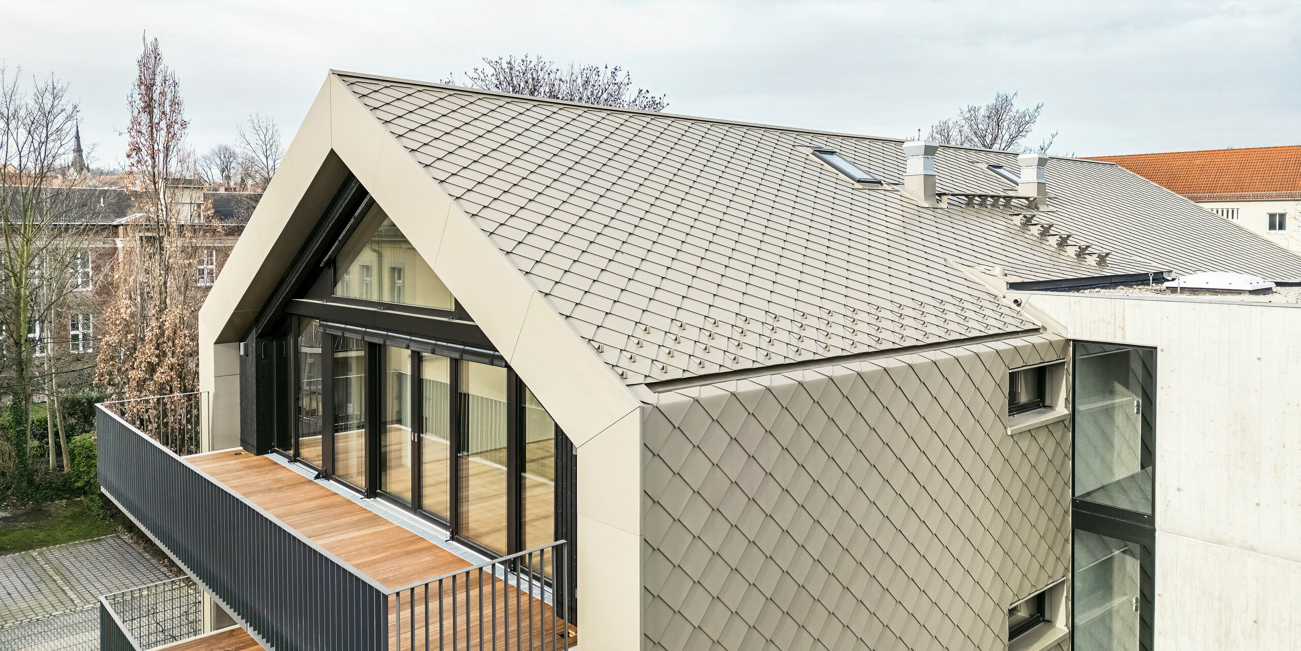 Vogelperspektive auf ein modernes Mehrfamilienhaus in der Chemnitzer Straße (Dresden) mit einer Gebäudehülle aus PREFA Aluminium-Rauten 29 × 29 in Bronze. Das Bild zeigt die elegante Dachstruktur, die großen Glasfronten und Balkone sowie die harmonische Integration des Gebäudes in die Umgebung. Die hochwertigen Materialien unterstreichen die langlebige Architektur des Gebäudes.