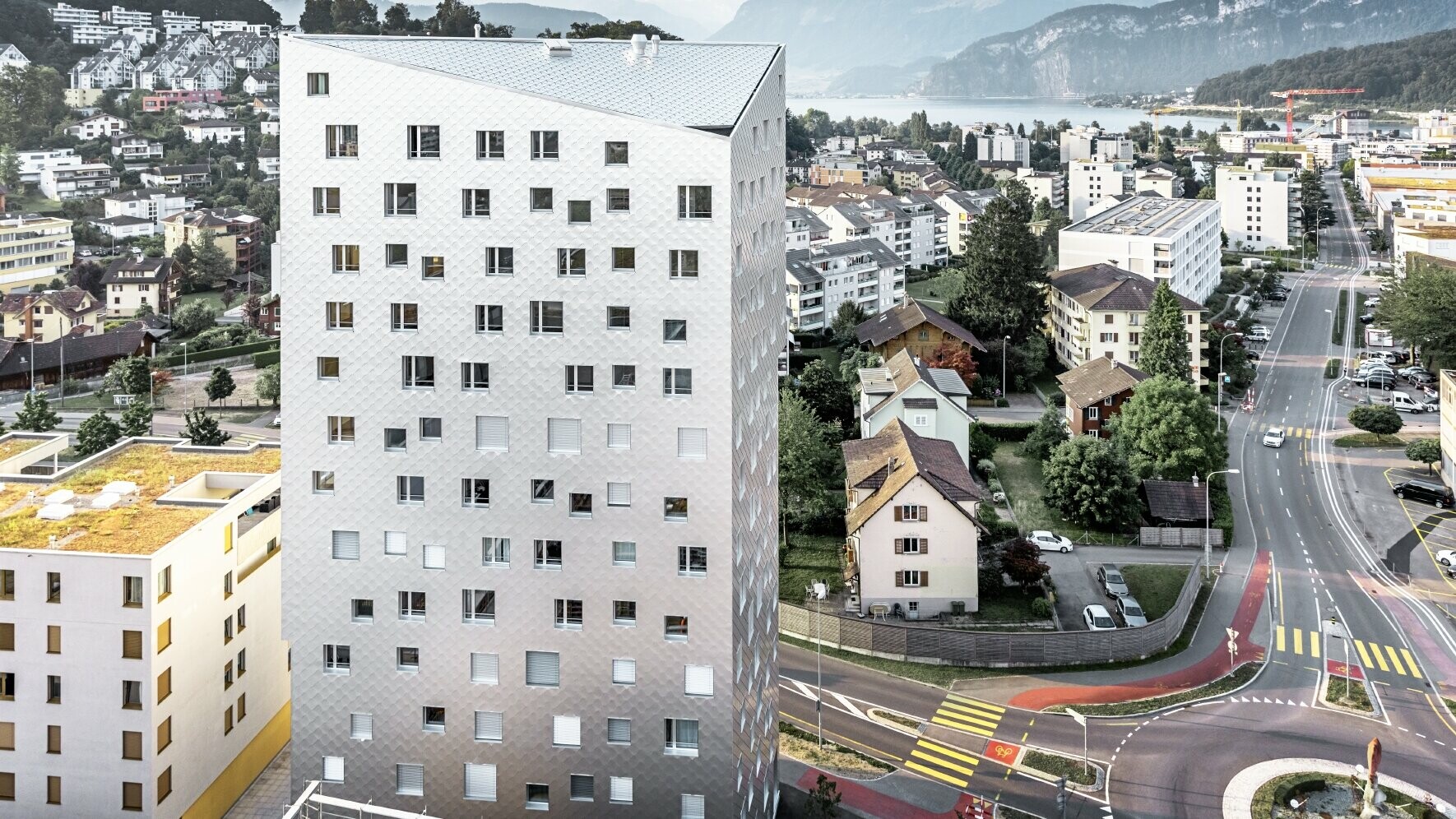 Das Bild zeigt das Solitaire-Hochhaus in Horw, einem Vorort von Luzern, das von der Schweizer Architektin Tilla Theus entworfen wurde. Das Hochhaus hat eine Fassade aus eloxierten Aluminiumrauten, die die Umgebung und die Wolken reflektieren. Die Fenster sind unregelmäßig angeordnet und als 'tanzende Fenster' bekannt. Im Hintergrund sind weitere Gebäude, Straßen und die malerische Berglandschaft der Schweiz zu sehen. Das Hochhaus steht inmitten einer städtischen Umgebung mit einer Mischung aus Wohnhäusern und städtischer Infrastruktur.