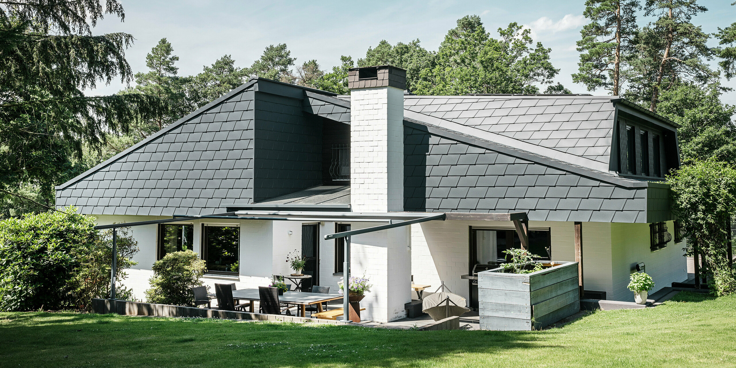 Vue arrière d'une maison individuelle à Mechernich avec terrasse et toit en bardeaux DS.19 PREFA en P.10 anthracite. Le toit en aluminium de haute qualité et la façade de l'étage supérieur sont résistants aux intempéries et de conception moderne. La façade en plâtre crépis crée un contraste élégant avec les produits en aluminium sombre, tandis que la terrasse est décorée avec goût avec des sièges et des plantes. Des arbres et des buissons encadrent la maison dans son environnement naturel.