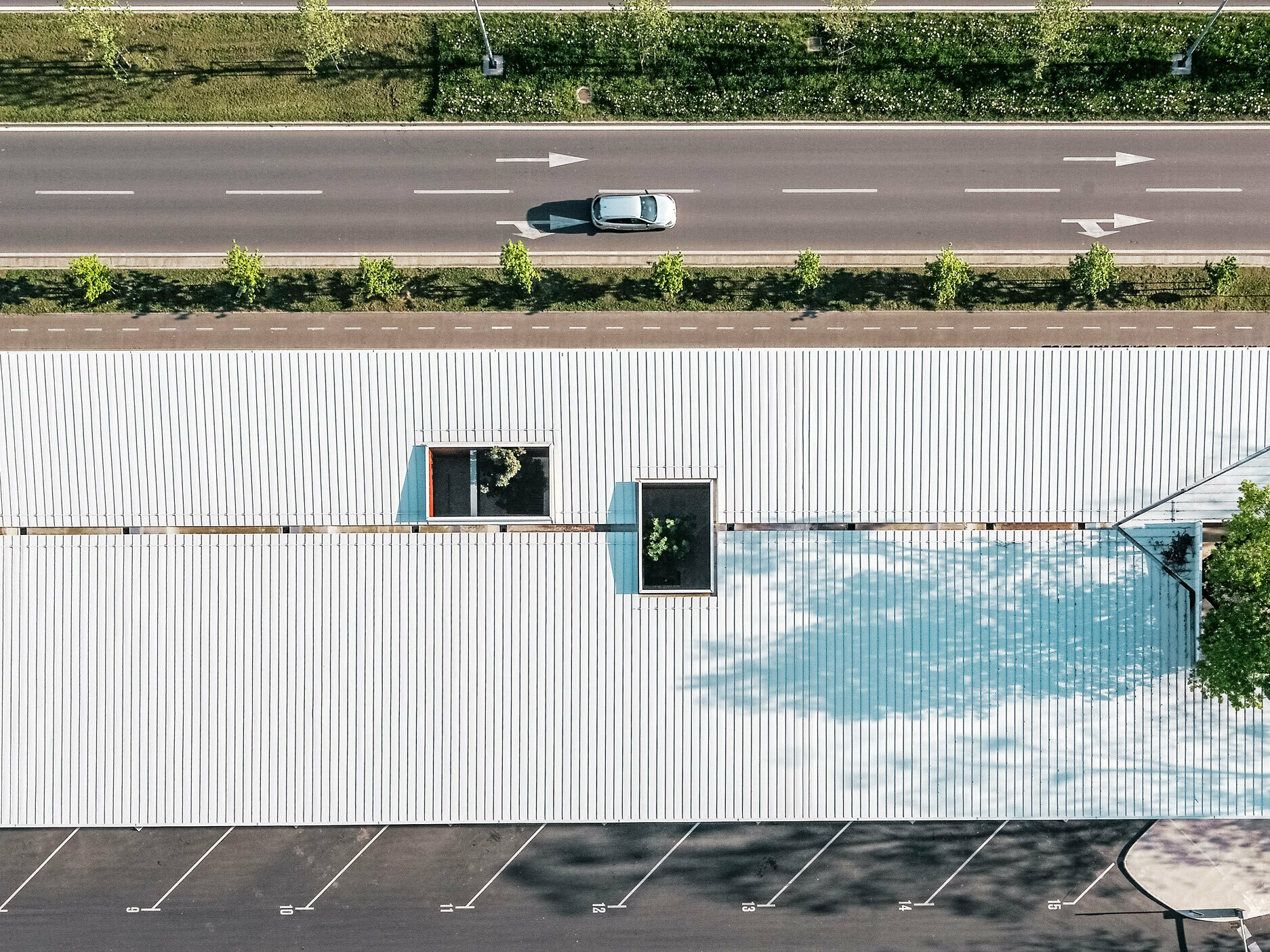 Das Bild zeigt eine Luftaufnahme der Bushaltestelle in Kroatien mit einem weißen Prefalz Dach von PREFA. Das lange Gebäude hat mehrere rechteckige Dachfenster, die in verschiedenen Positionen angeordnet sind und das grüne Laub von Bäumen durchscheinen lassen. Ein kleinerer Parkplatz ist im Vordergrund zu sehen, während sich im Hintergrund eine zweispurige Straße mit einer Linie von Bäumen entlangzieht. 