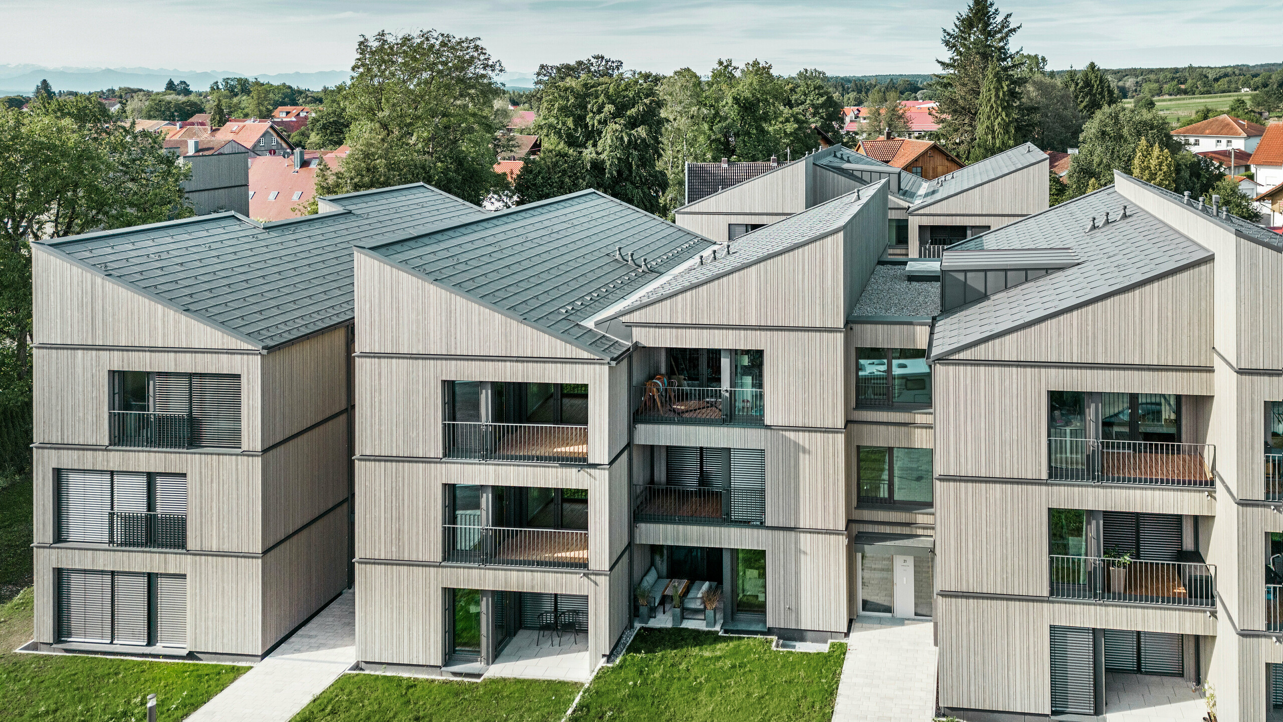 Vista aerea del moderno condominio sul sito Schmucker di Utting am Ammersee, con costruzione ibrida in legno sostenibile ed eleganti facciate in legno. Gli edifici sono dotati di tegole PREFA R.16 in P.10 grigio scuro. Questa architettura innovativa, progettata dallo studio WWA Architekten di Monaco, è caratterizzata da linee chiare e da un'armoniosa integrazione nel verde circostante. Gli ampi balconi e le grandi finestre creano un'atmosfera abitativa luminosa e accogliente, mentre i materiali di alta qualità garantiscono durata e resistenza agli agenti atmosferici.