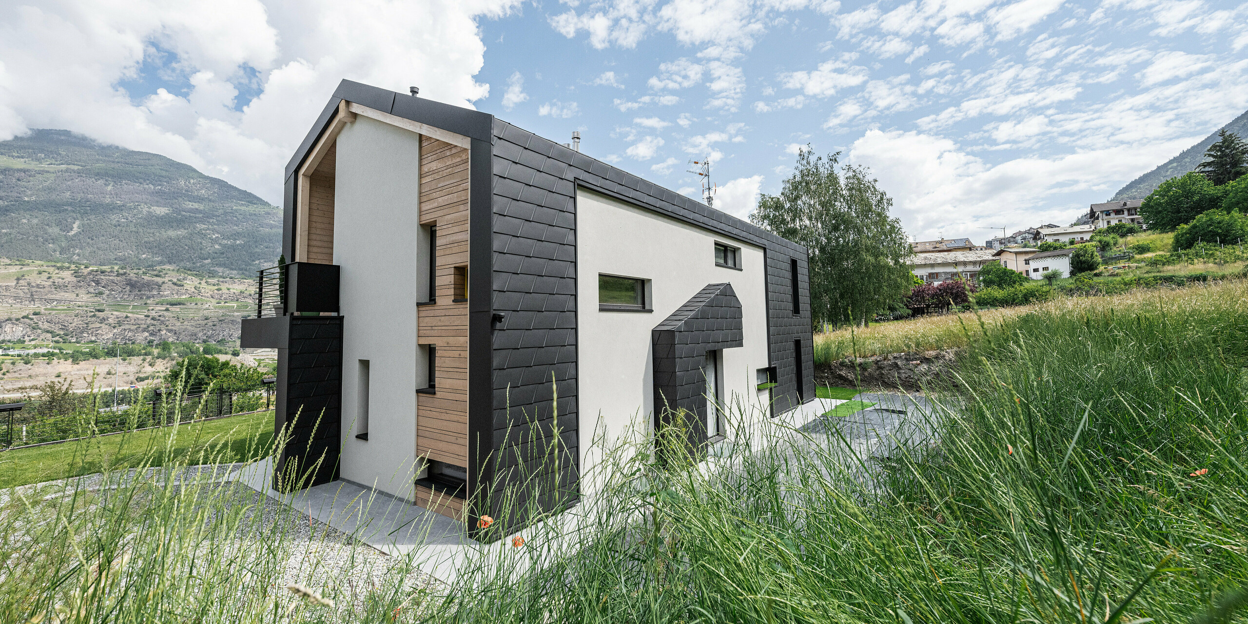 Ein Einfamilienhaus in Italien mit schwarzen PREFA Dachschindeln DS.19 und Wandschindeln, eingebettet in eine idyllische Landschaft mit Blick auf bewaldete Hügel. Die Schindeln aus Aluminium bieten eine moderne, langlebige Außenverkleidung, die natürliche Holzelemente ergänzt und für eine stilvolle Optik sorgt. Die Gräser im Vordergrund und die klare Bergluft vermitteln ein Gefühl von Ruhe und Naturverbundenheit.