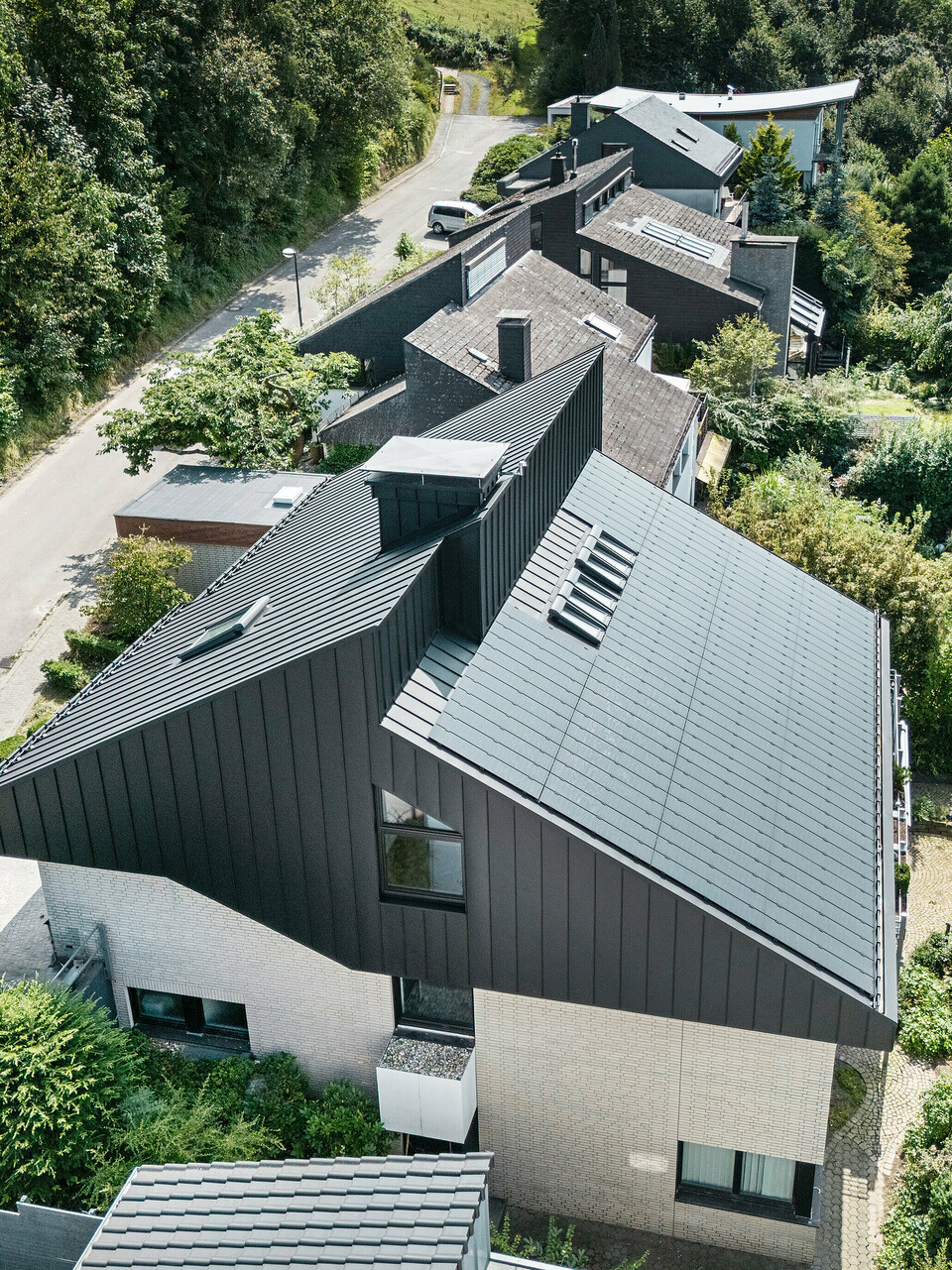 Vue oblique d'une maison unifamiliale moderne à Meschede, en Allemagne. Le toit en pente décalé a été revêtu de PREFALZ en P.10 noir. Le toit en aluminium constitue un élément visuel marquant et offre au bâtiment une protection permanente. La toiture à double joint debout de précision s'étend du toit à travers la façade jusqu'aux pignons. Entouré d'une végétation luxuriante et d'une rue calme, le bâtiment économe en énergie avec les modules solaires innovants PREFALZ s'intègre parfaitement dans l'environnement de vie naturel.