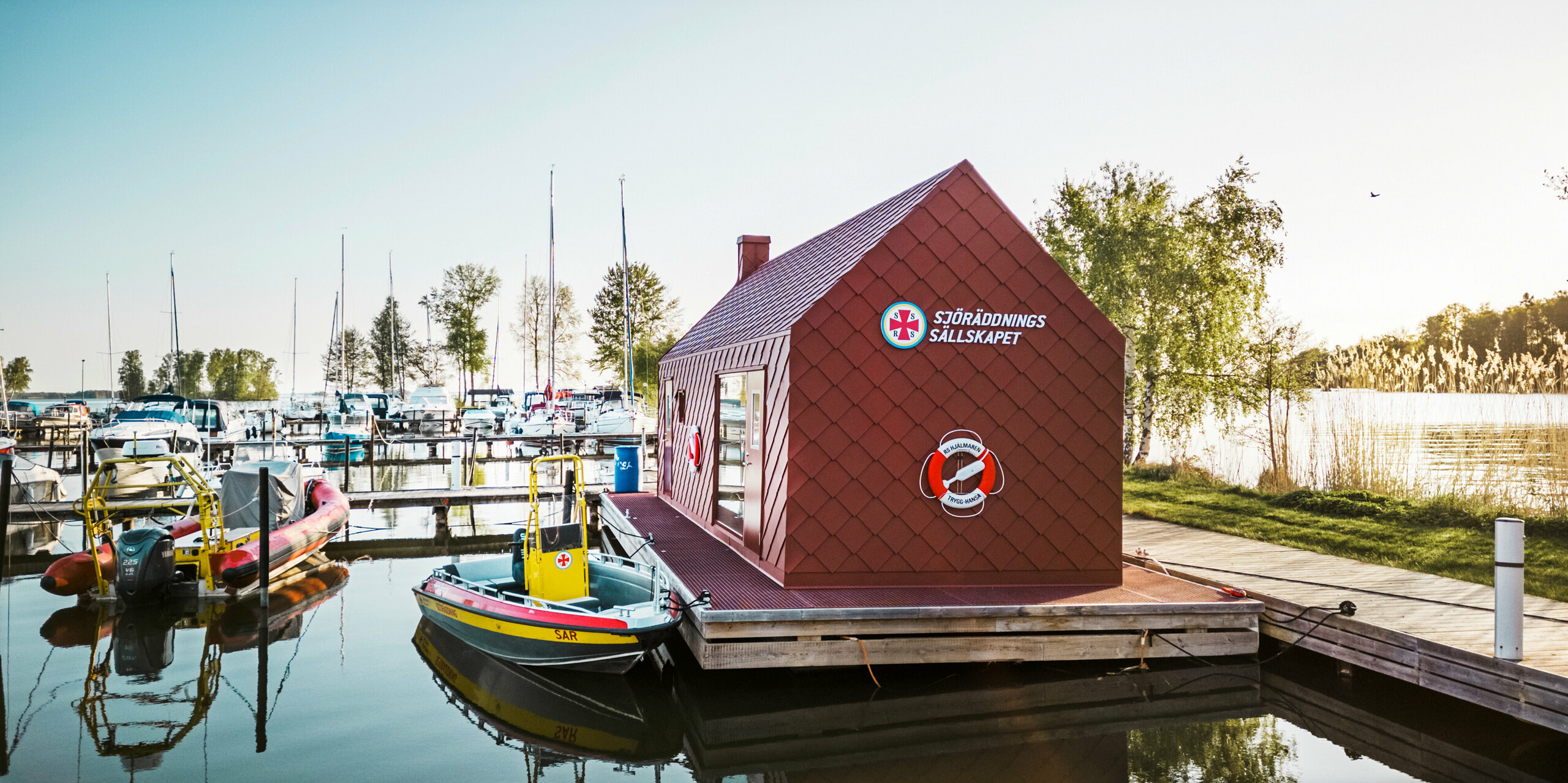 Morgenstimmung mit der schwimmenden Rettungsstation am Hjälmaren See, Schwedens viertgrößtem See. Das Wasserhaus mit seiner auffälligen oxydroten PREFA Dach- und Wandraute 29×29 Verkleidung steht im Vordergrund und hebt sich deutlich von der Umwelt ab. Rettungsboote liegen bereit am Steg, während im Hintergrund die Segelboote im ruhigen Hafenbecken spiegeln. Die Szene vermittelt eine Atmosphäre der Ruhe und Sicherheit, unterstützt durch die prägnante Architektur und Farbgebung der Station, die als Ankerpunkt für die Seenotrettung dient.