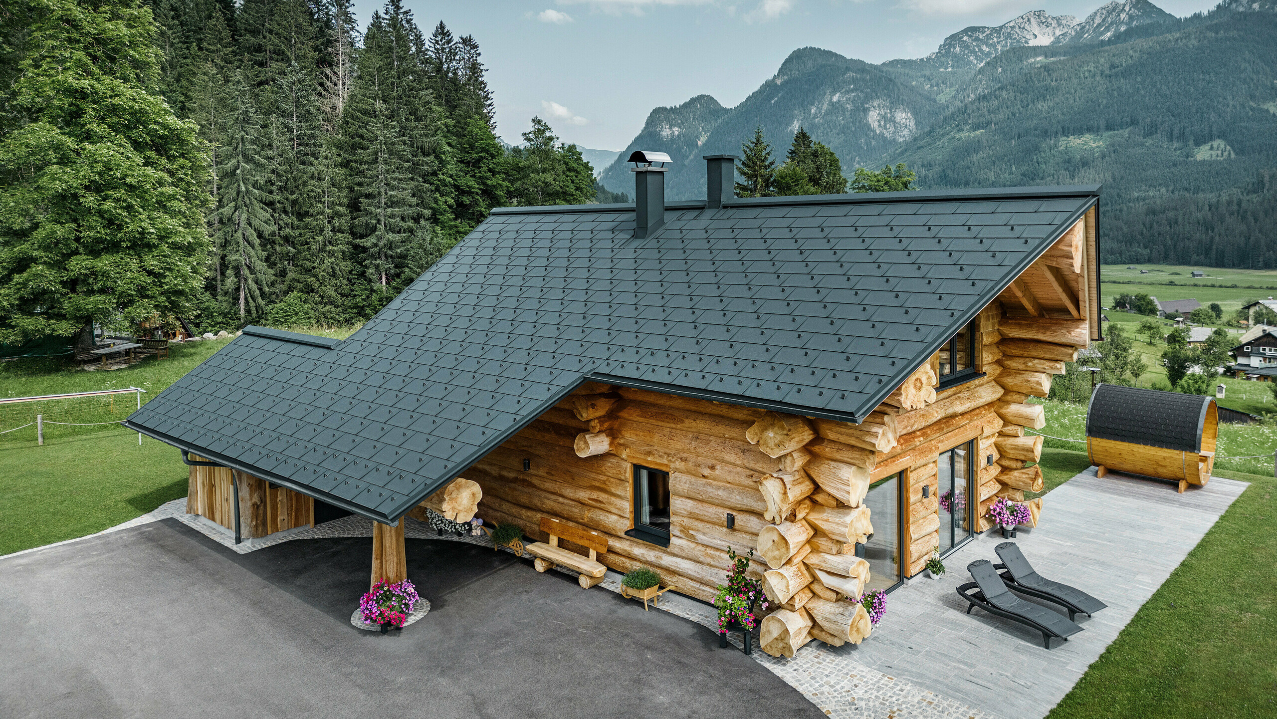 Schräge Ansicht eines malerischen Blockhauses in Gosau, Österreich, ausgestattet mit einem PREFA Dach aus anthrazitfarbenen Dachplatten R.16. Die Architektur kombiniert rustikales Blockhausflair mit modernen Elementen, umgeben von dichten grünen Wäldern und einer beeindruckenden Bergkulisse. Das Haus strahlt mit seiner ländlichen Eleganz und den präzise verlegten Dachelementen aus Aluminium, die für ihre Langlebigkeit und Witterungsbeständigkeit bekannt sind, eine einladende Atmosphäre aus.