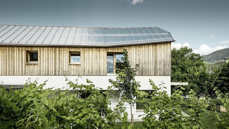 Seitliche Ansicht des Einfamilienhauses in Pruggern eingedeckt mit PREFALZ in der Farbe Naturblank. Die Hälfte des Hauses ist durch eine Hecke verdeckt. Das Dach schimmert leicht im Sonnenschein.