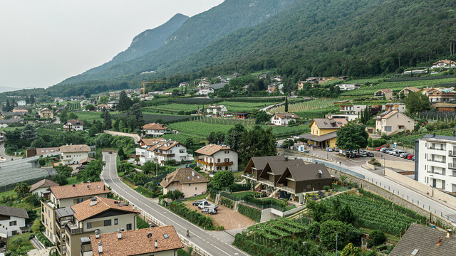 Drohnenaufnahme eines Dorfes in einem bergigen Gebiet in Südtirol. Im Zentrum des Bildes stehen drei Reihenhäuser, hervorgehoben durch eine markante Aluminiumhülle aus PREFA Dach- und Wandrauten 44x44 in der Farbe P.10 Braun. Die moderne Architektur fügt sich nahtlos in die traditionelle Umgebung ein, umgeben von üppigen grünen Weinbergen und dichten Wäldern.