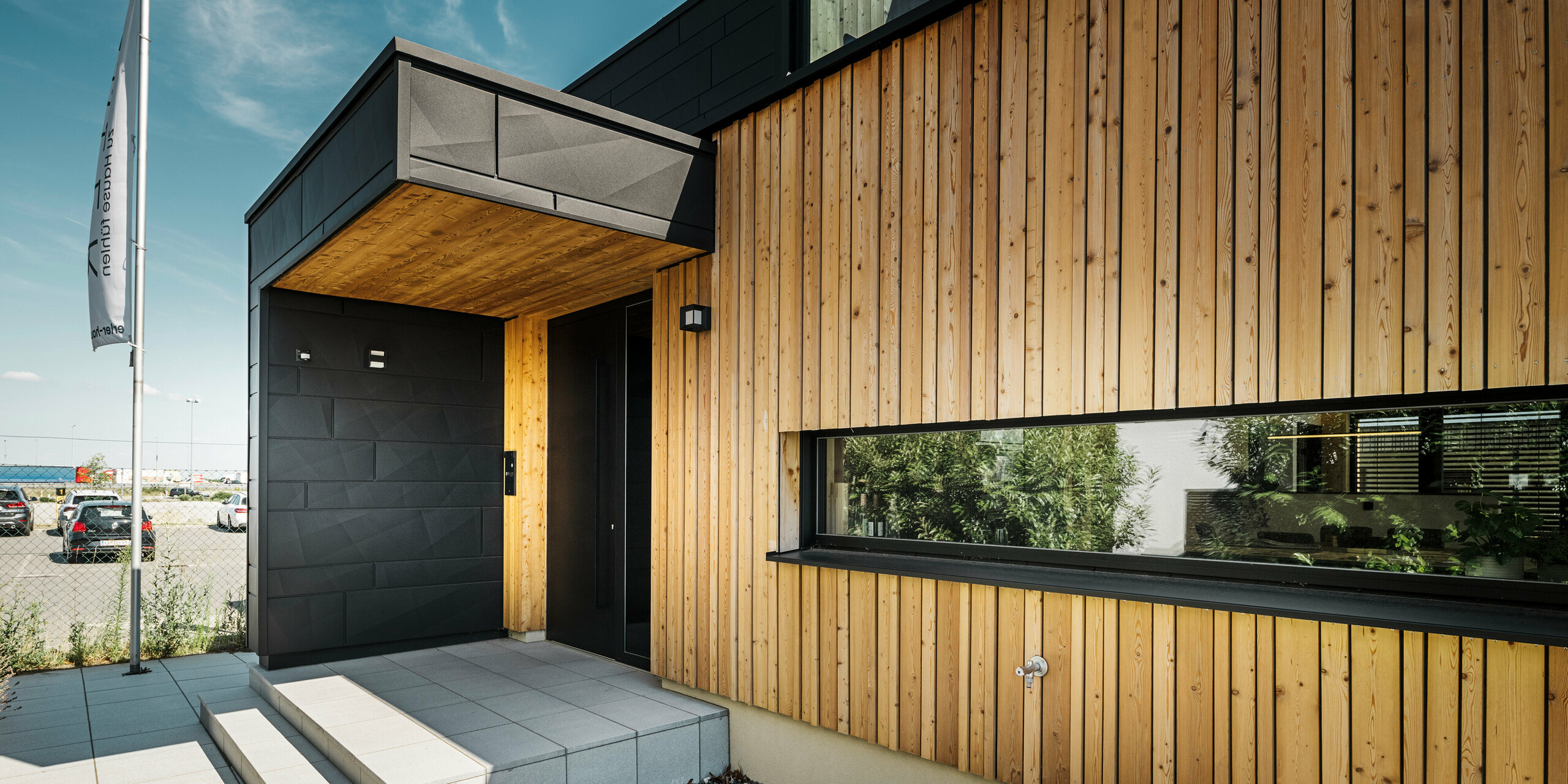 Entrée latérale de la maison modèle Autark du centre de maisons préfabriquées Blaue Lagune à Vösendorf, avec une façade moderne en bois naturel et des panneaux PREFA Siding.X noirs. L'image montre la combinaison élégante du revêtement en bois et de l'aluminium dans le revêtement de haute qualité P.10 noir.