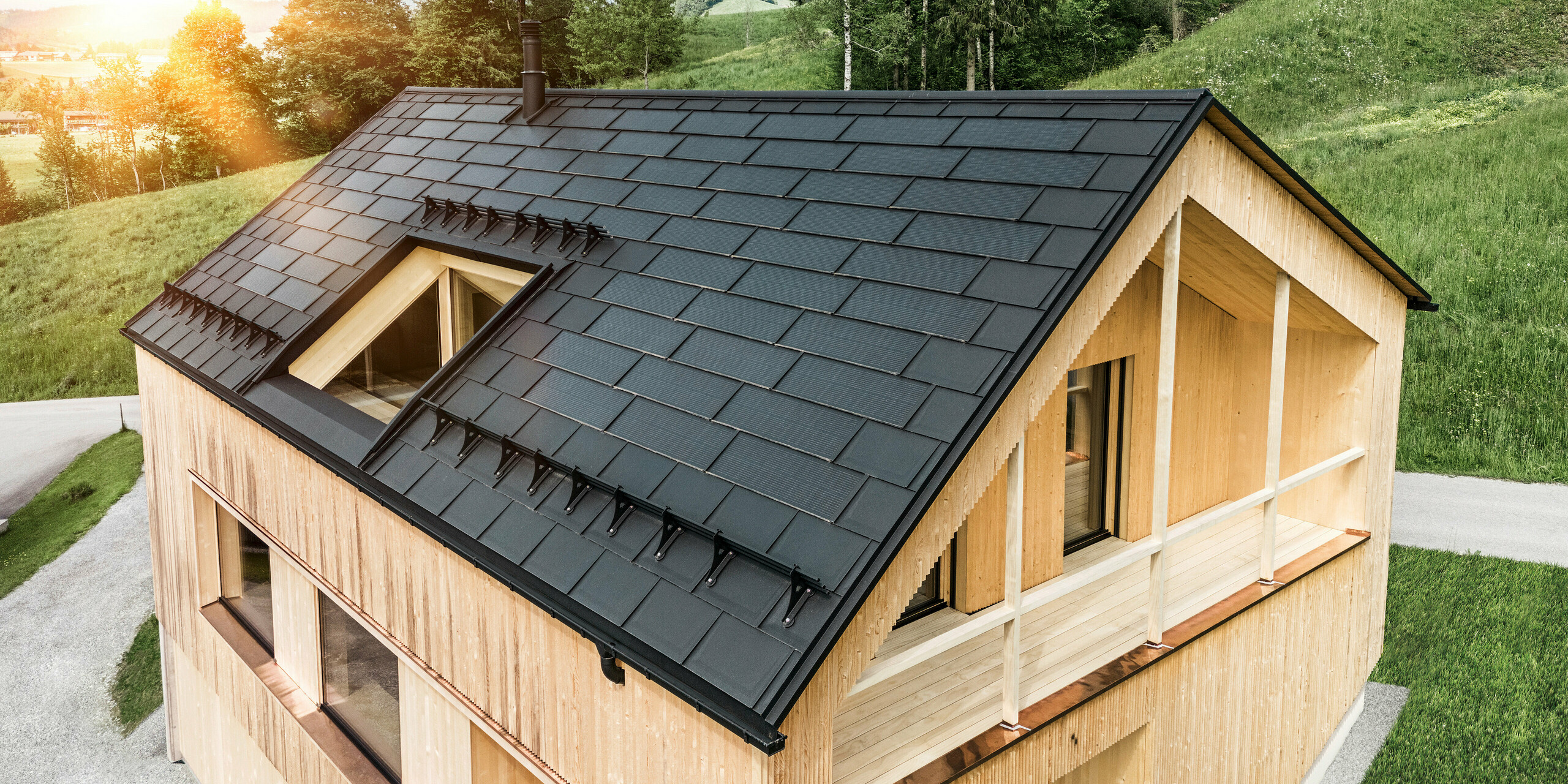 Prise de vue d'une maison en bois moderne et durable à Egg avec un toit solaire PREFA, qui se distingue par son aspect sombre, structuré et élégant. Les modules solaires sont stratégiquement placés entre les R.16 classiques pour profiter d'un rayonnement solaire optimal et s'intégrer esthétiquement dans la conception globale. La maison est en harmonie avec son environnement verdoyant, tandis que le chaud soleil du soir met en valeur la texture naturelle du bois de la façade et souligne la solidité de la construction. Un exemple harmonieux de bâtiment respectueux de l’environnement et d’efficacité énergétique.