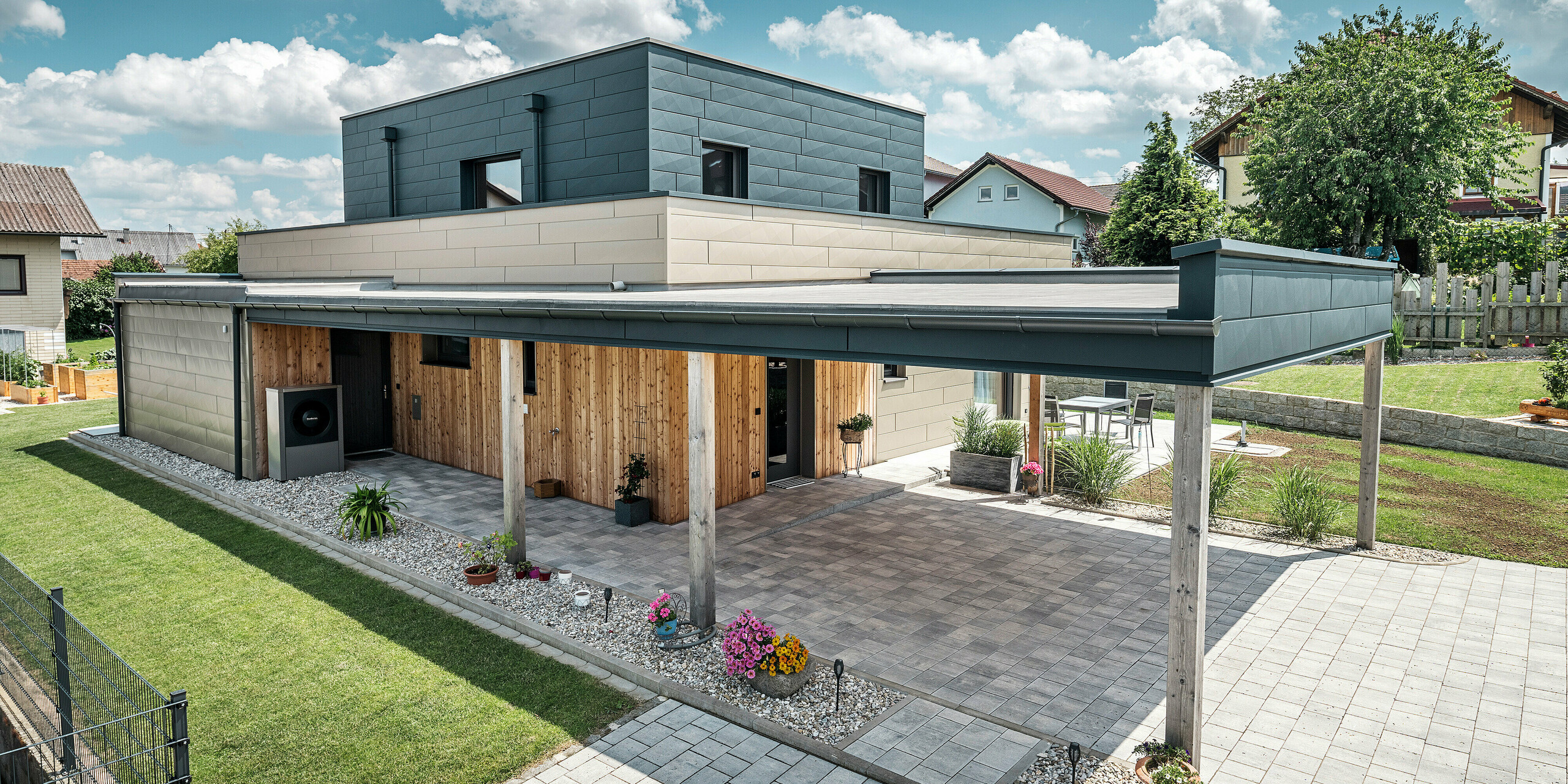 Luftaufnahme der Zufahrt eines modernen Einfamilienhauses in Wippenham, Österreich, mit einer zweifarbigen PREFA Aluminiumfassade in Bronze und Anthrazit. Das Haus zeigt die stilvolle Integration von Siding.X Fassadenpaneelen und Holzverkleidung. Deutlich sichtbar ist der überdachte Carport, der nahtlos in die Architektur des Hauses integriert ist. Der gepflegte Garten und die Terrassenfläche unterstreichen das moderne und funktionale Design des Gebäudes.